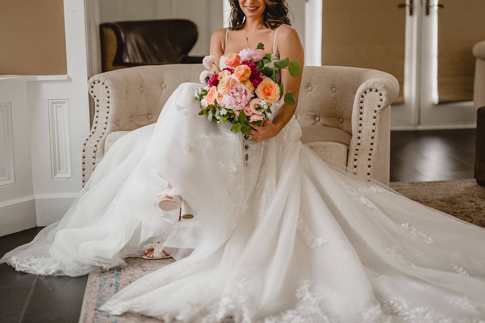 bride sits on tan couch holding bouquet of pink flowers
