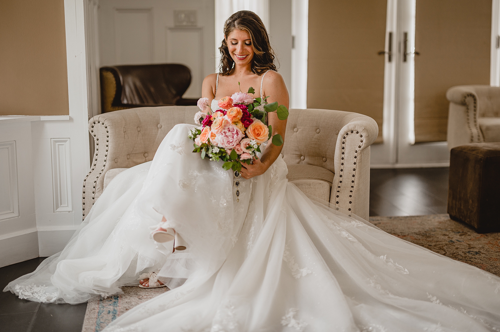 bride in wedding dress sits on tan couch holding bouquet of pink and red flowers