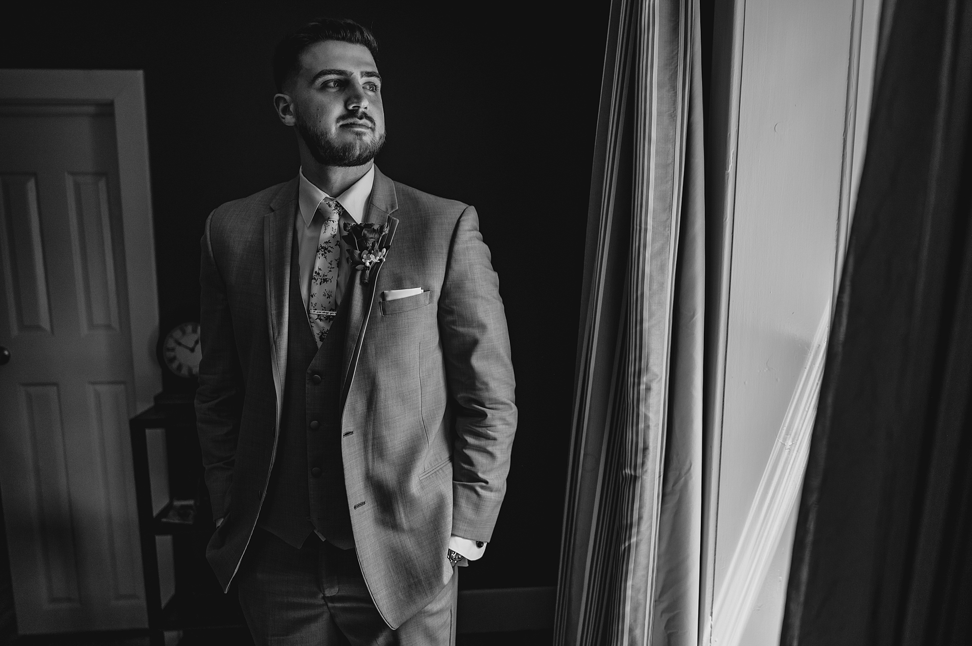 groom in light suit puts hands in pockets and looks out window