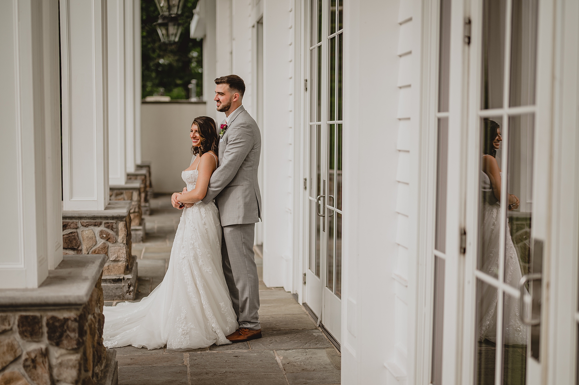 groom in grey suit hugs bride from behind in front of windows at the Ryland Inn