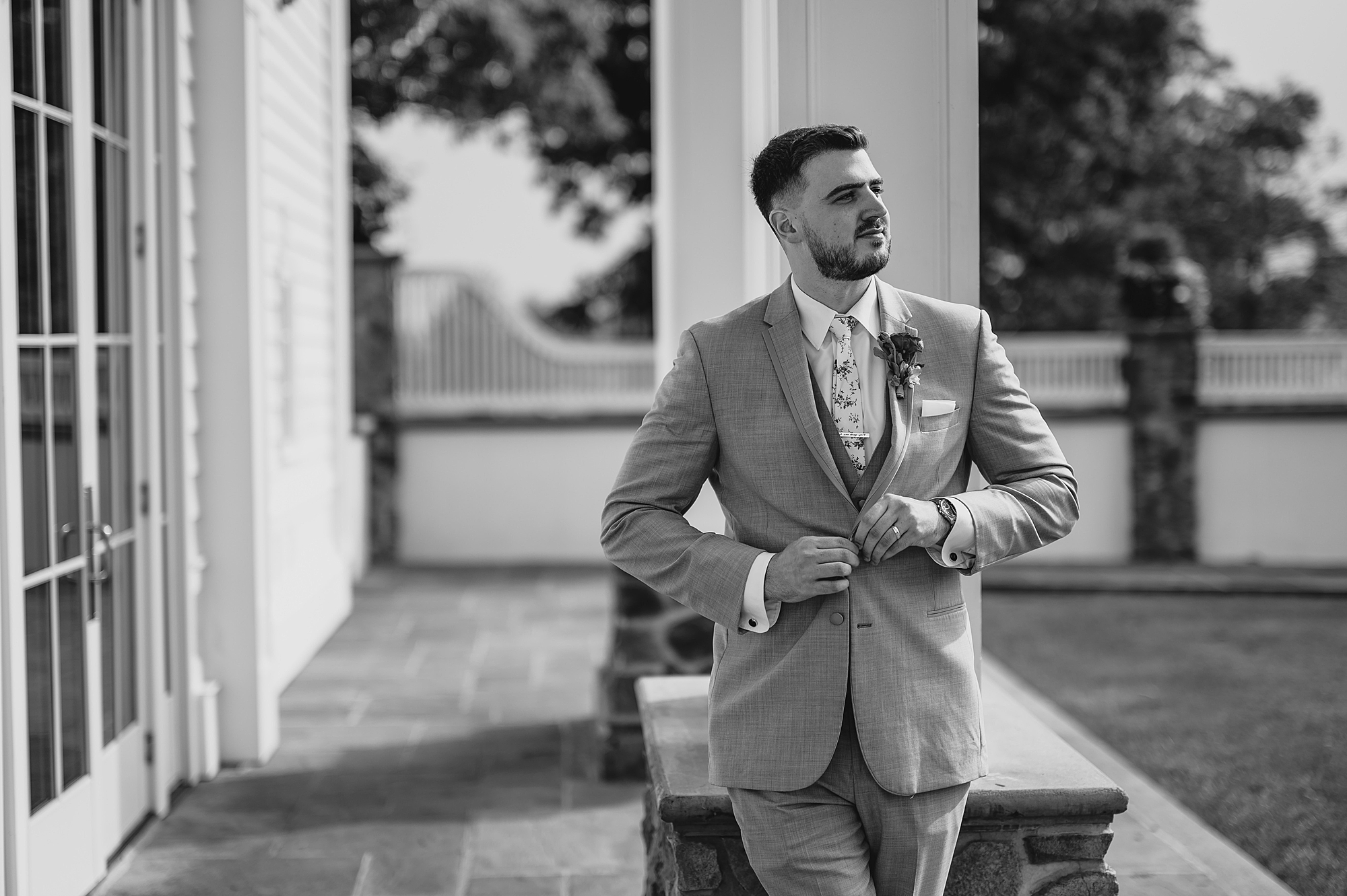 groom leans against pillar buttoning up suit jacket