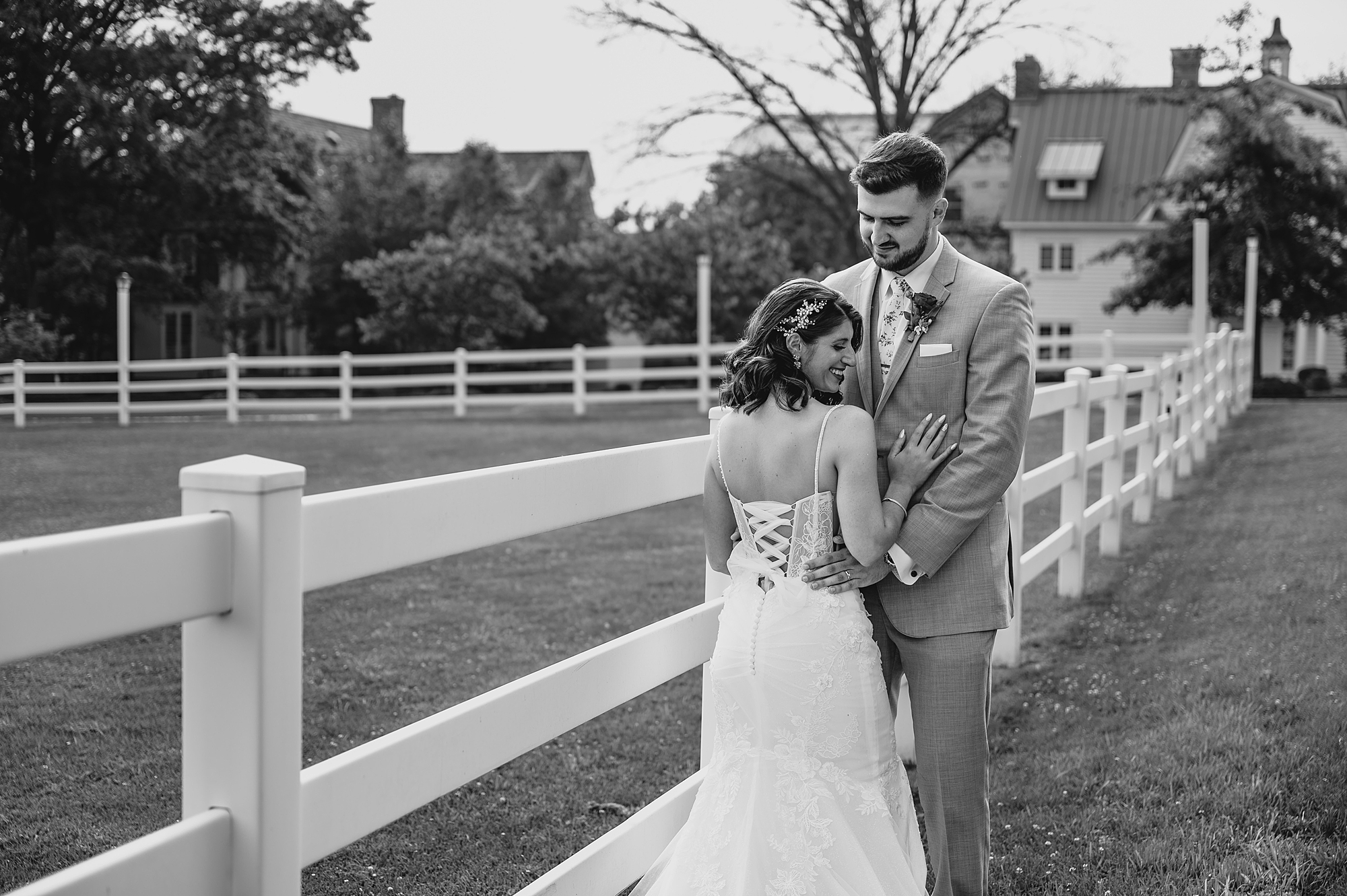 groom hugs bride looking down at her as they lean on fence