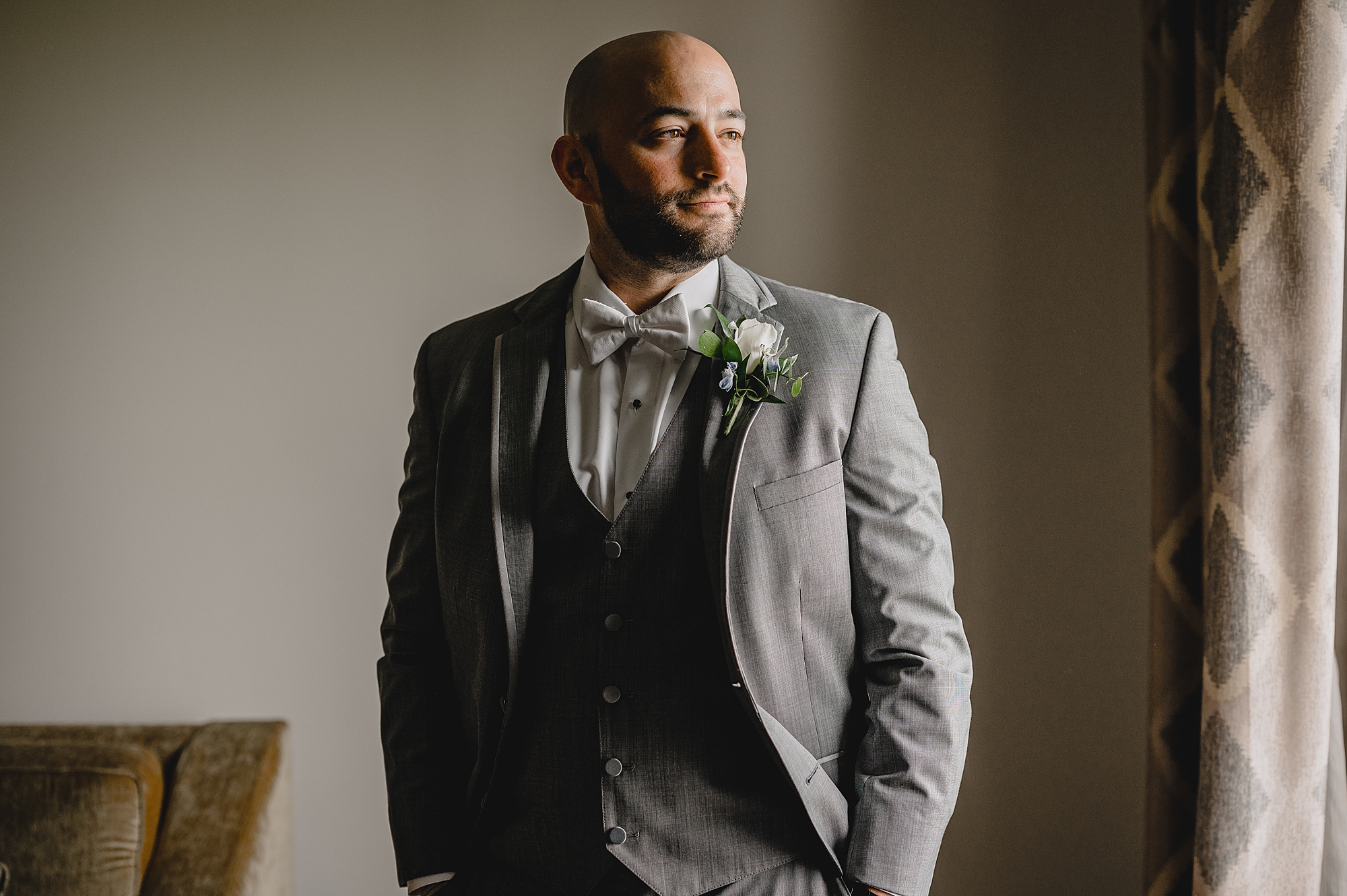 groom stands with hands in pockets of grey suit looking out window