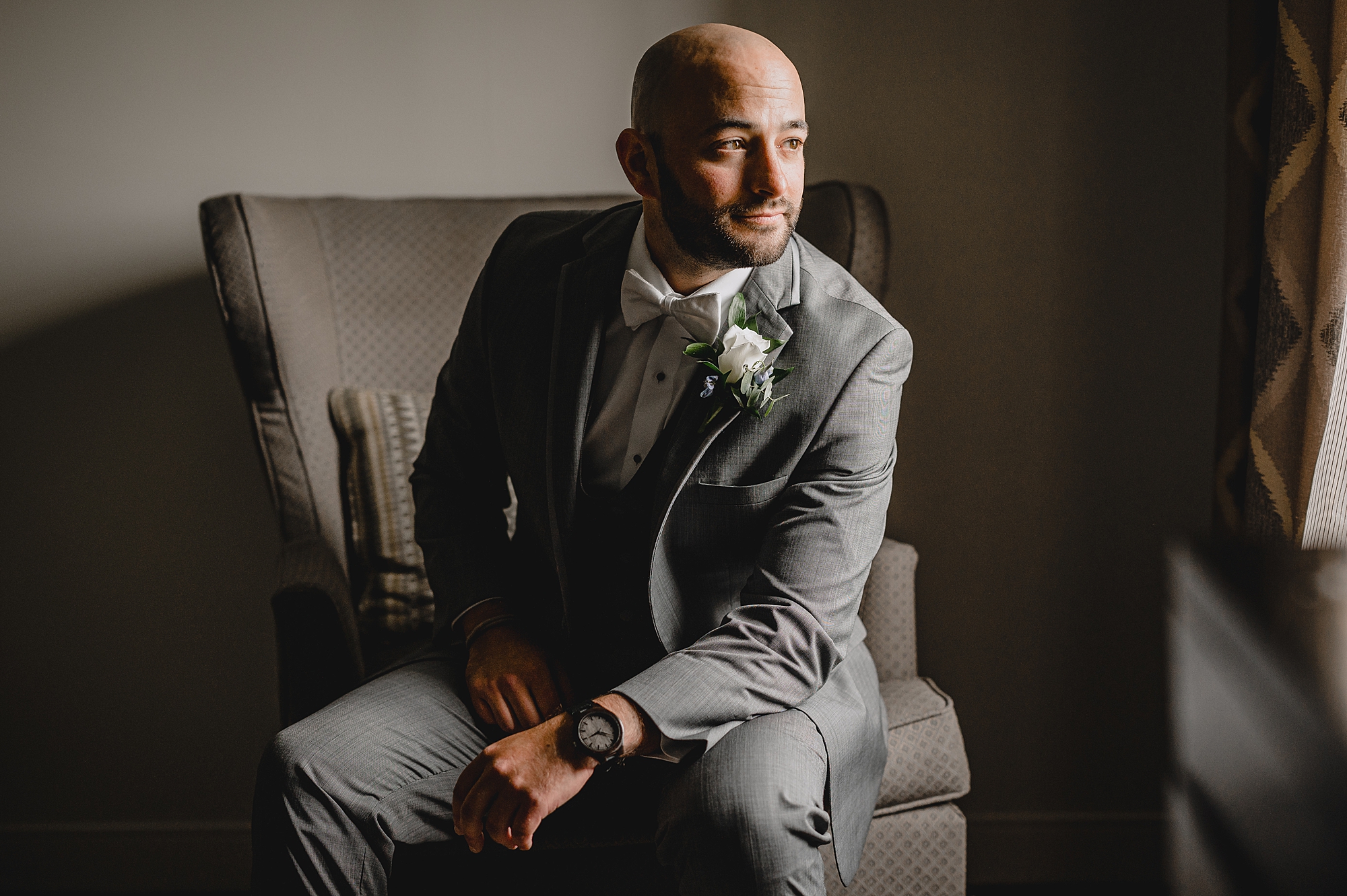groom sits in chair leaning forward on knee in grey suit