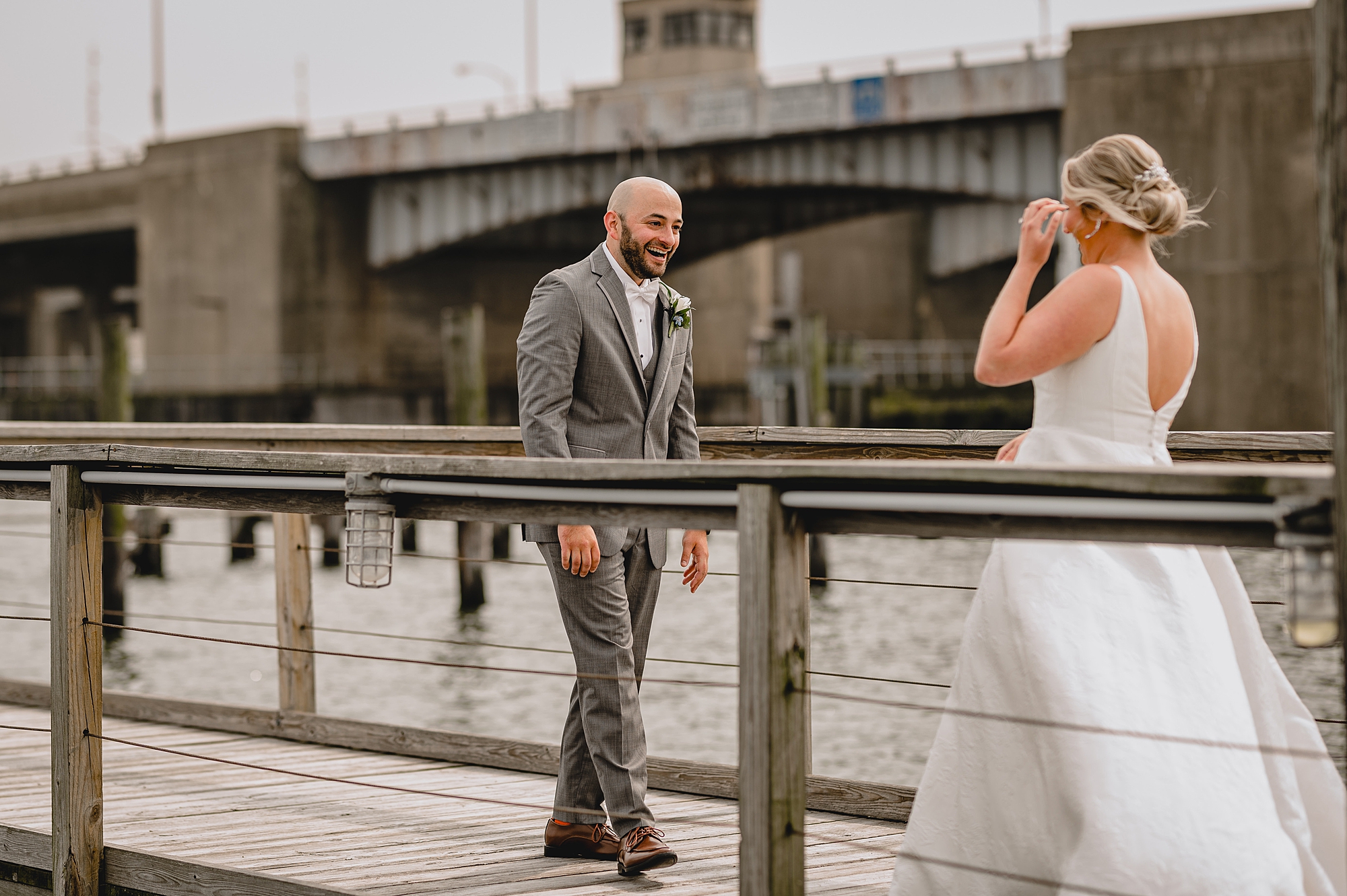 groom grins seeing bride in wedding dress on wooden bridge