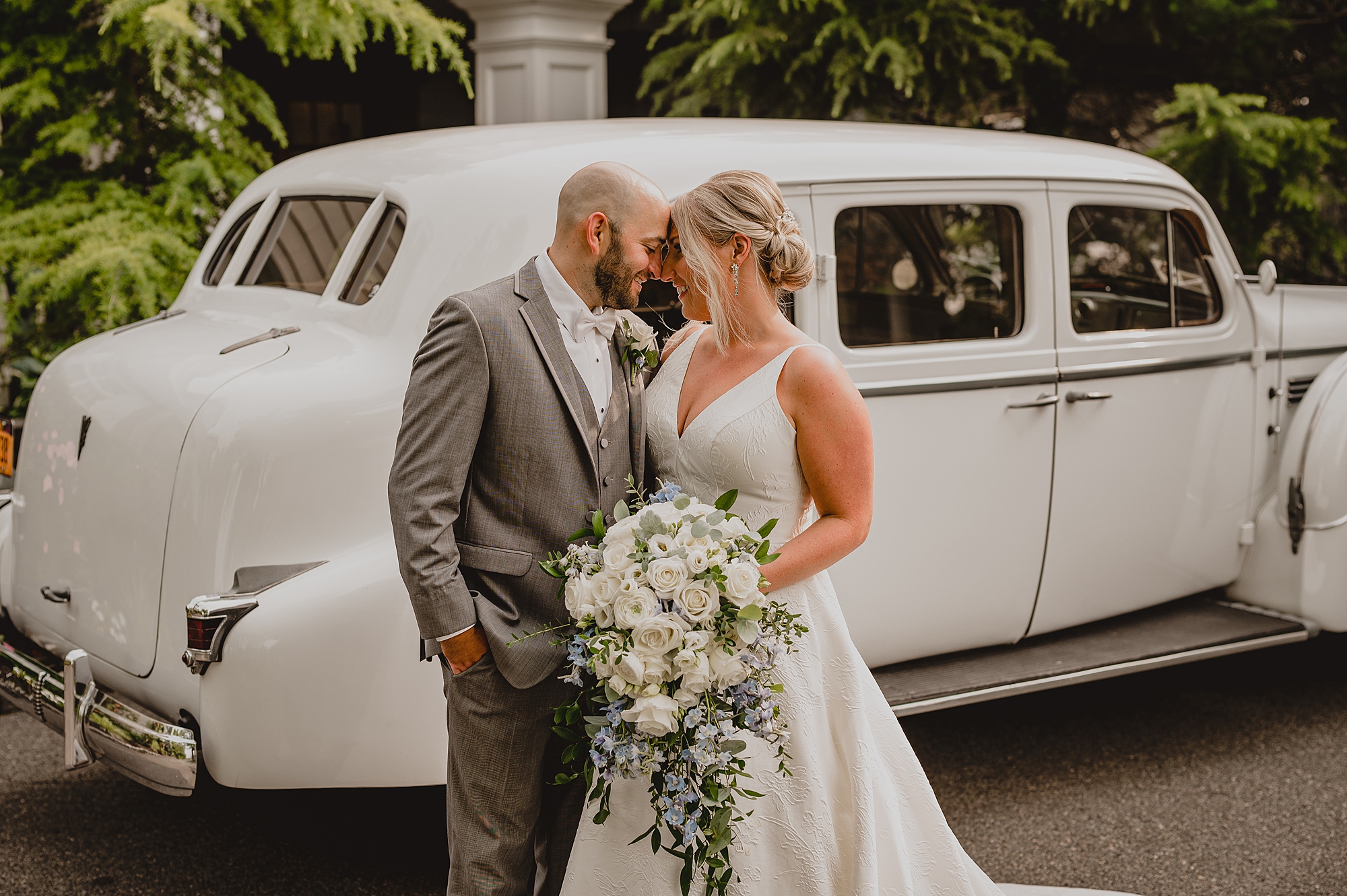 bride and groom lean heads together posing by classic white car