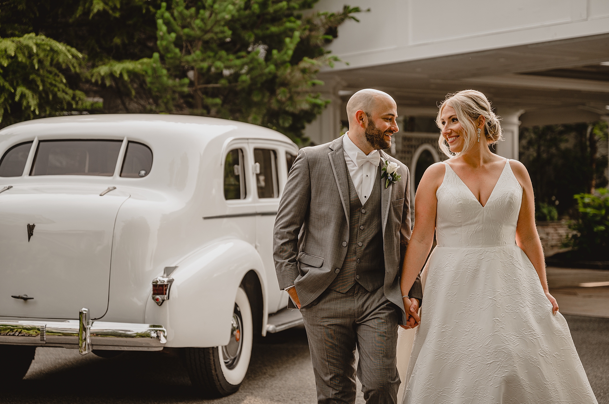 newlyweds smile holding hands walking by white vintage car