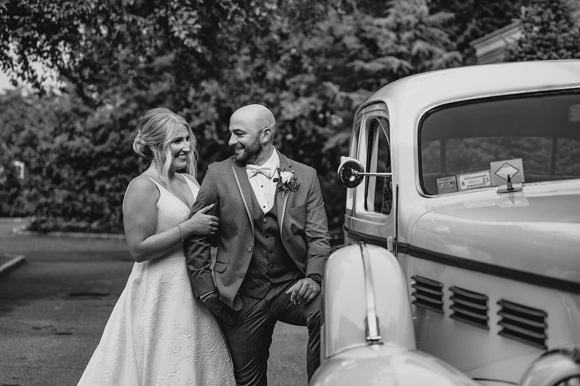 bride holds groom's arm while he poses against running board of white vintage car