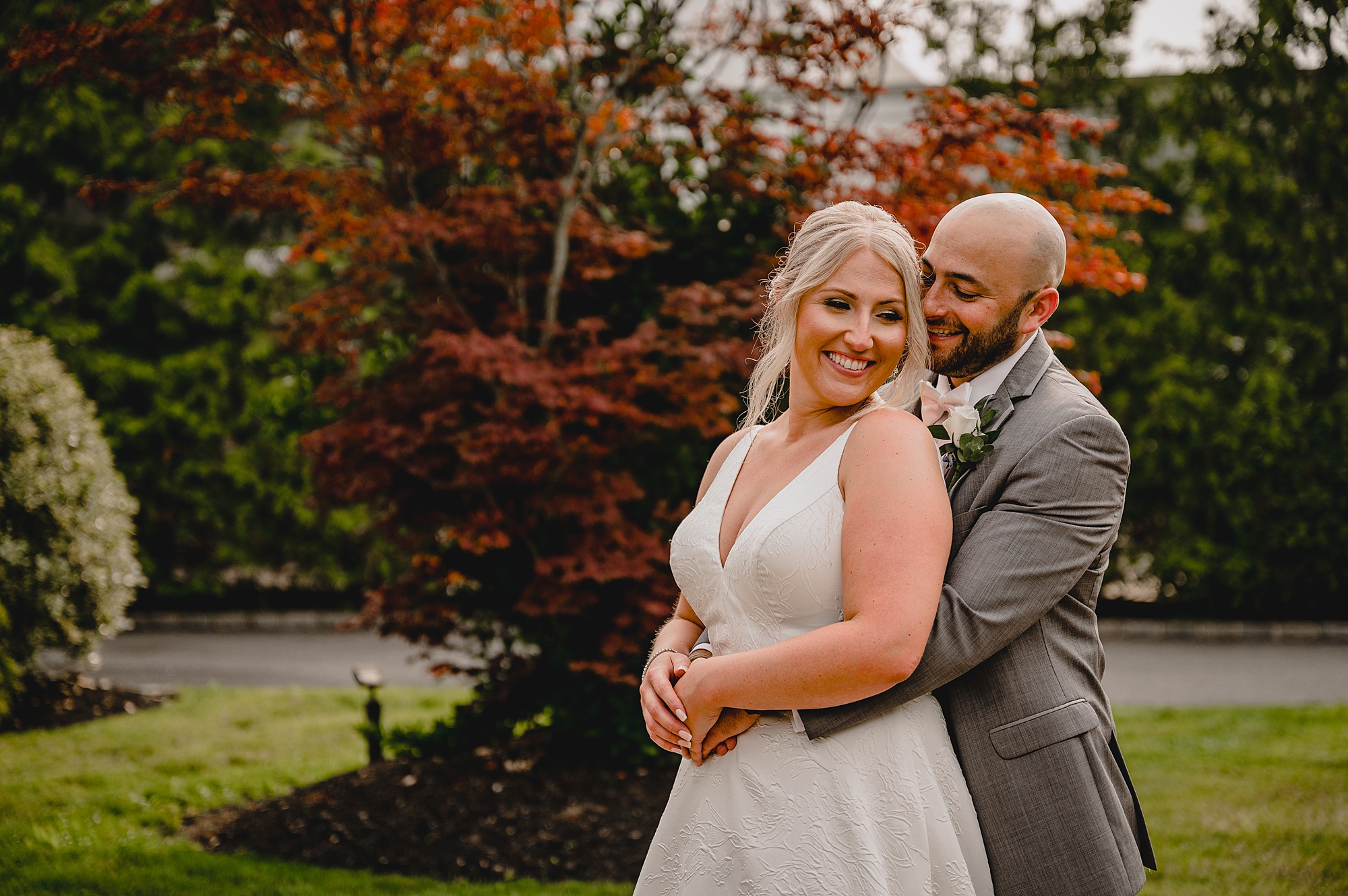 groom hugs bride from behind standing by tree with orange and red leaves