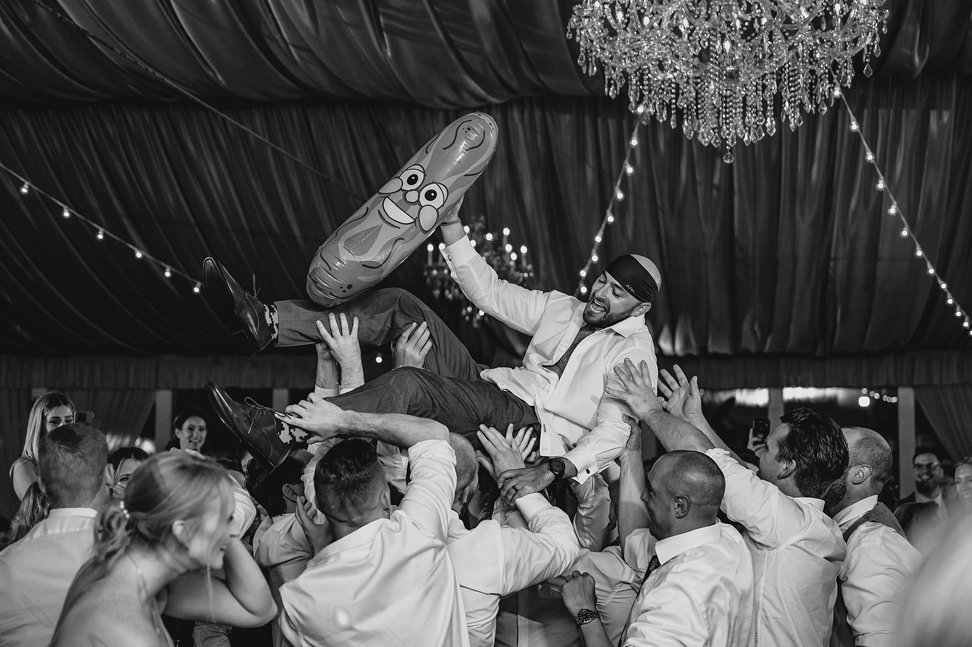 groom holds up inflated pickle while groomsmen hold him on dance floor