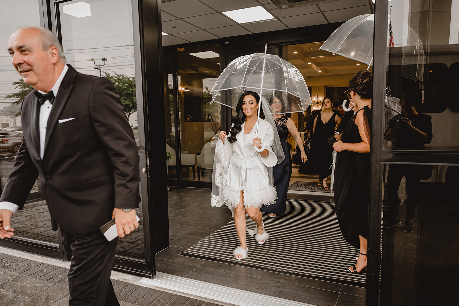 bride walks into hotel wearing white robe and carrying clear umbrella