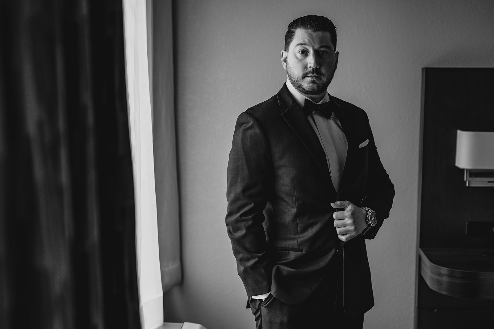 groom looks towards camera holding lapel of suit by bed in hotel suite