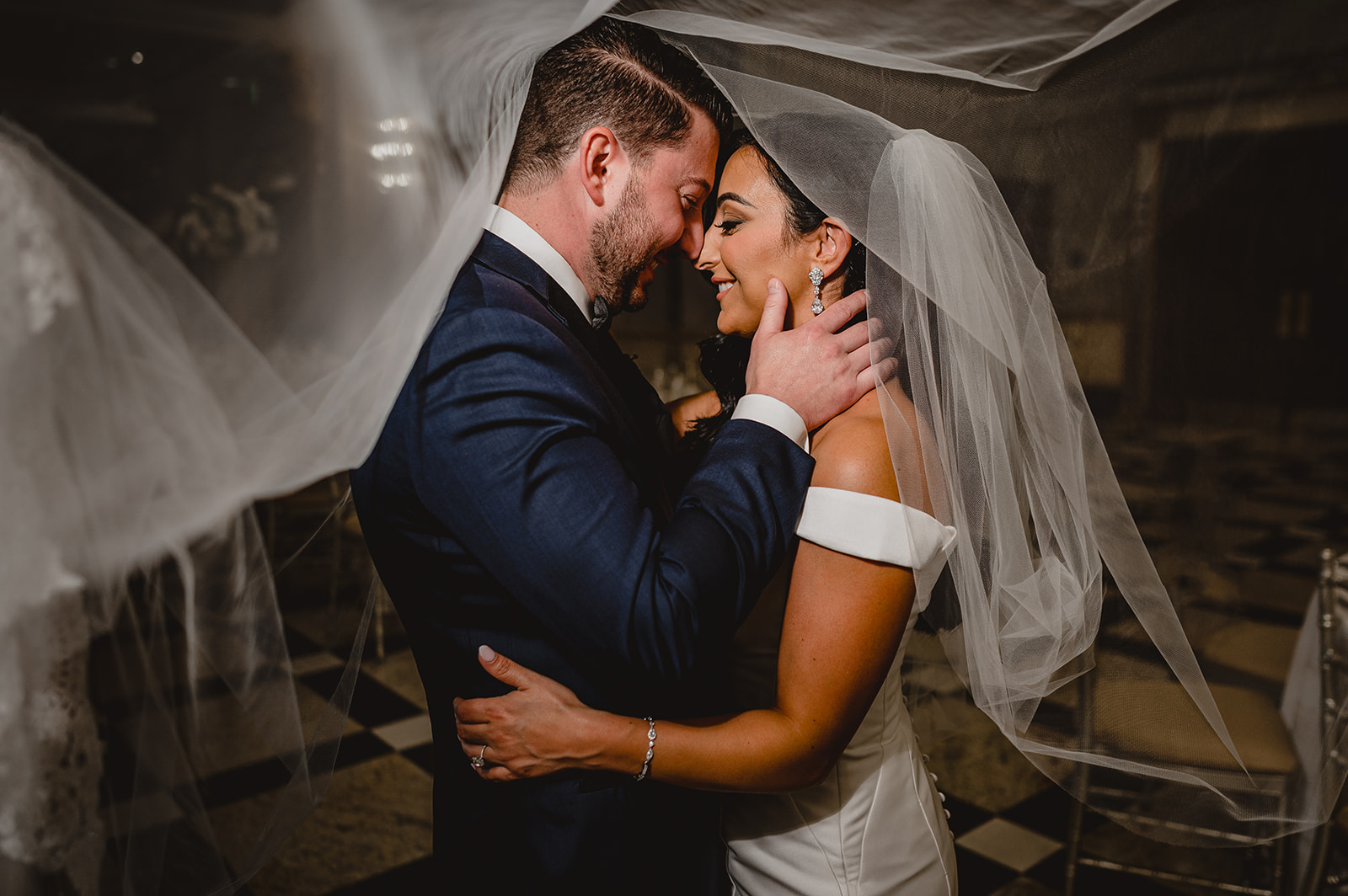 groom holds bride's chin under veil leaning to kiss her