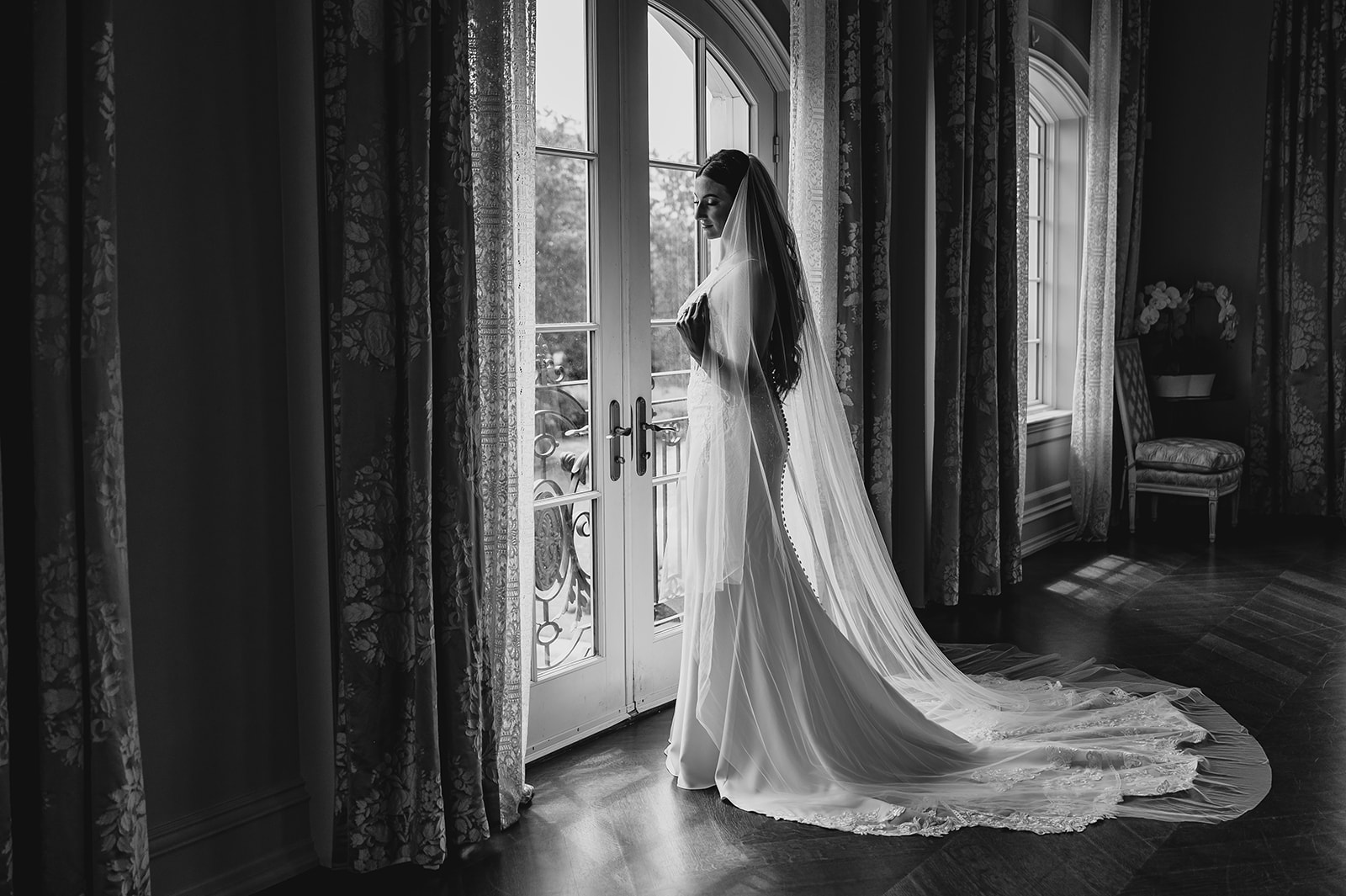 bride looks out window at Park Chateau Estate with wedding gown and veil laid out behind her