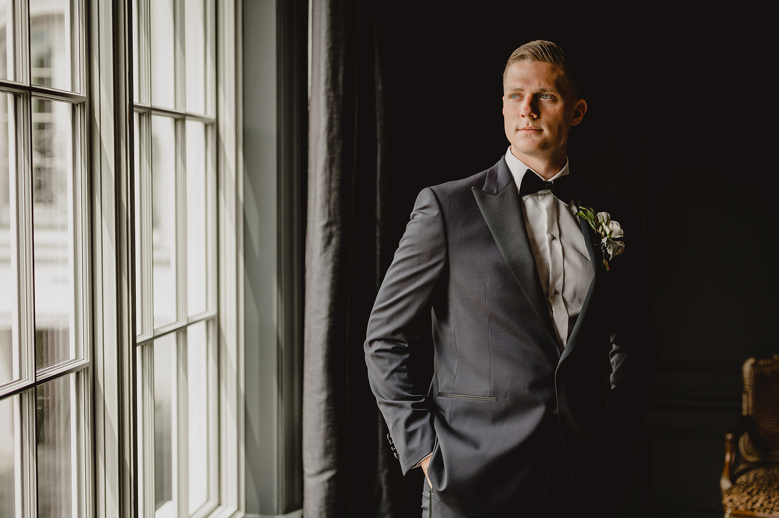 groom stands with hands in pockets of navy suit looking out window