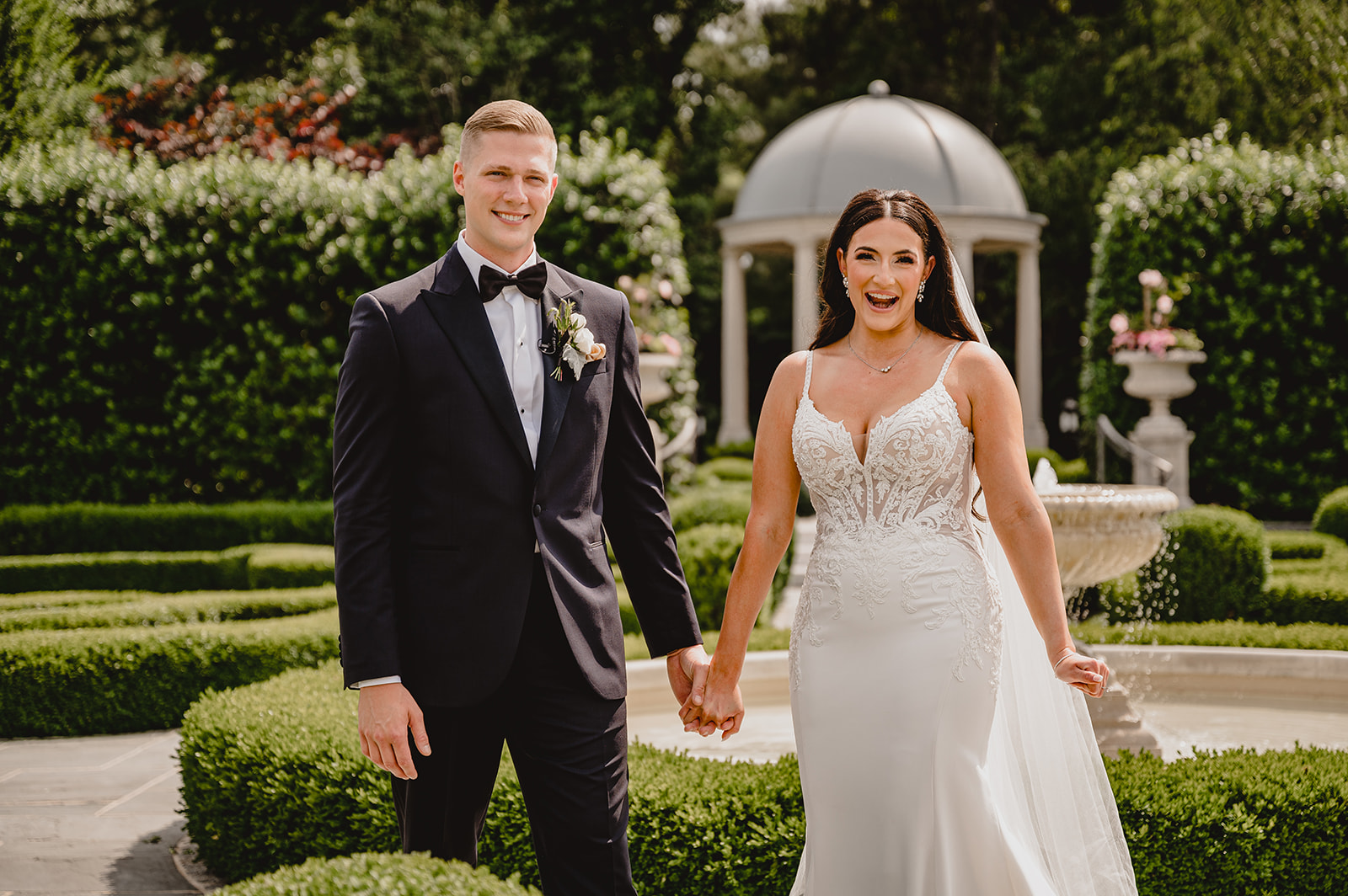 bride and groom hold hands between hedges and gazebo