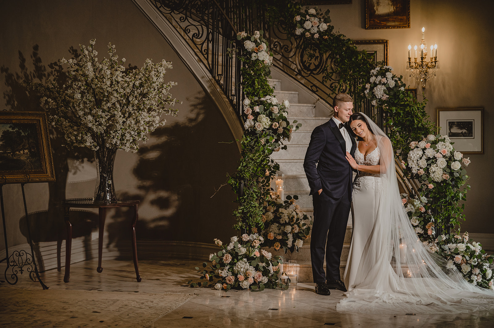 bride leans head on groom's chest at bottom of flower covered staircase inside Park Chateau Estate