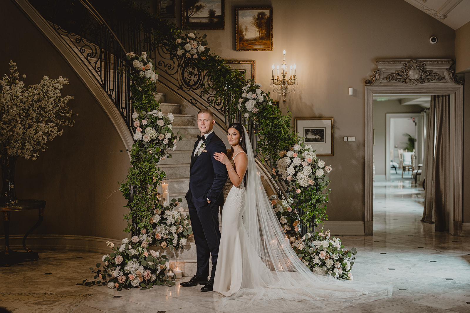bride hugs groom from behind at bottom of flower covered staircase inside Park Chateau Estate