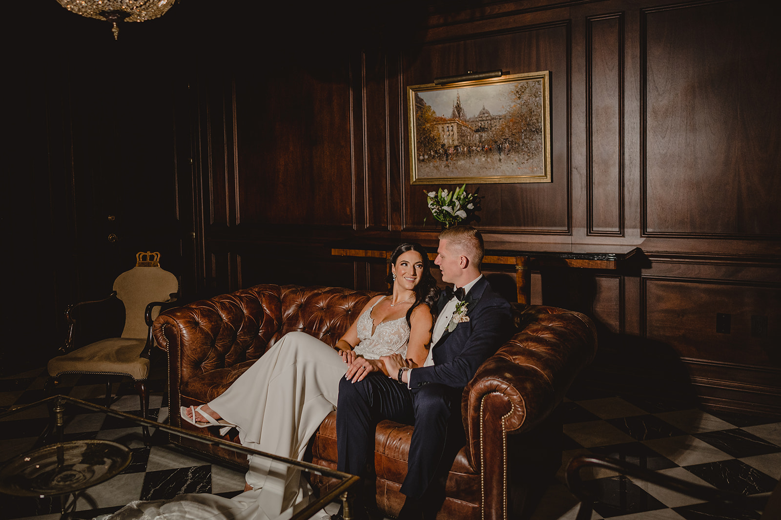 newlyweds sit on leather couch inside library at Park Chateau Estate