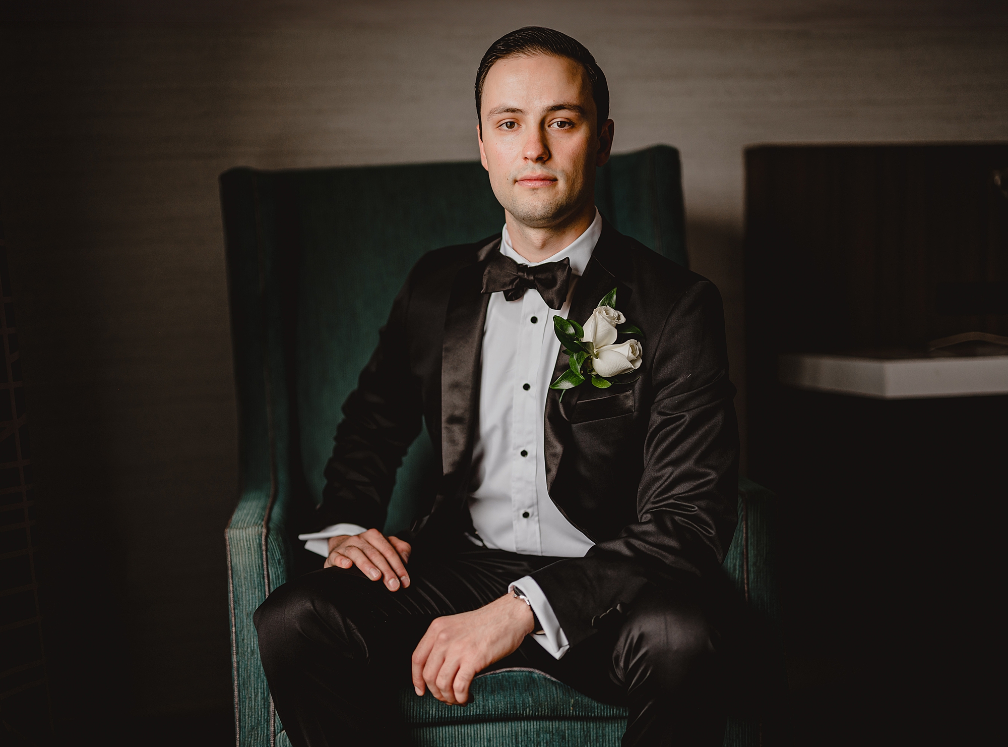 groom sits in green chair in black suit 