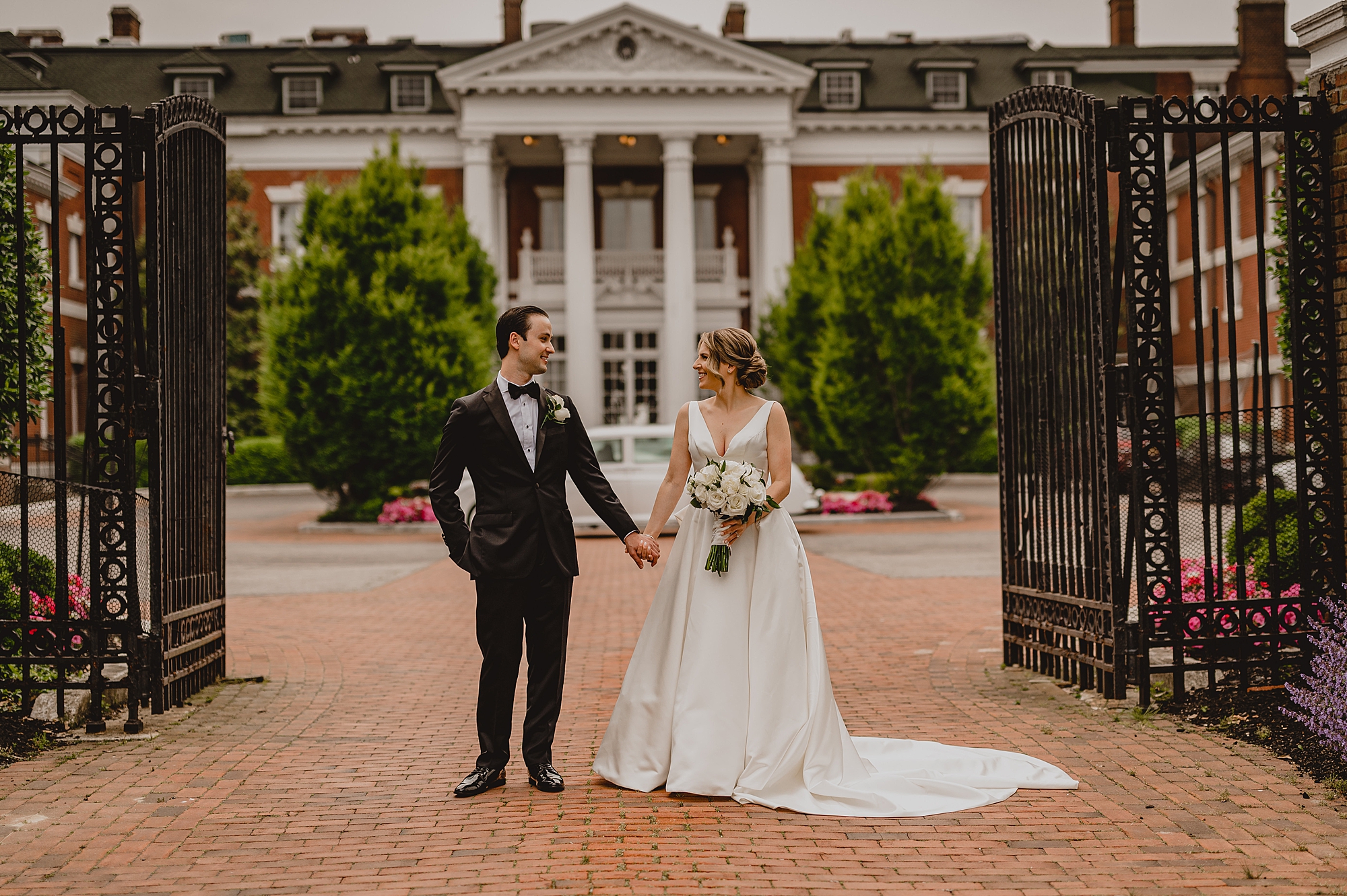 bride and groom hold hands standing between iron gates at Bourne Mansion