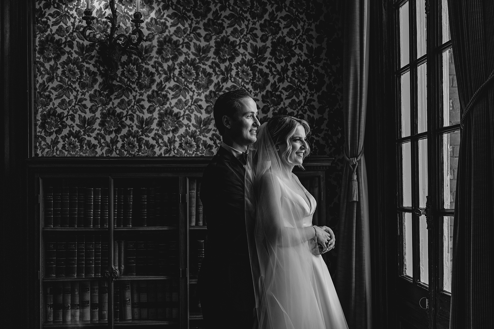 groom hugs bride from behind in front of window 