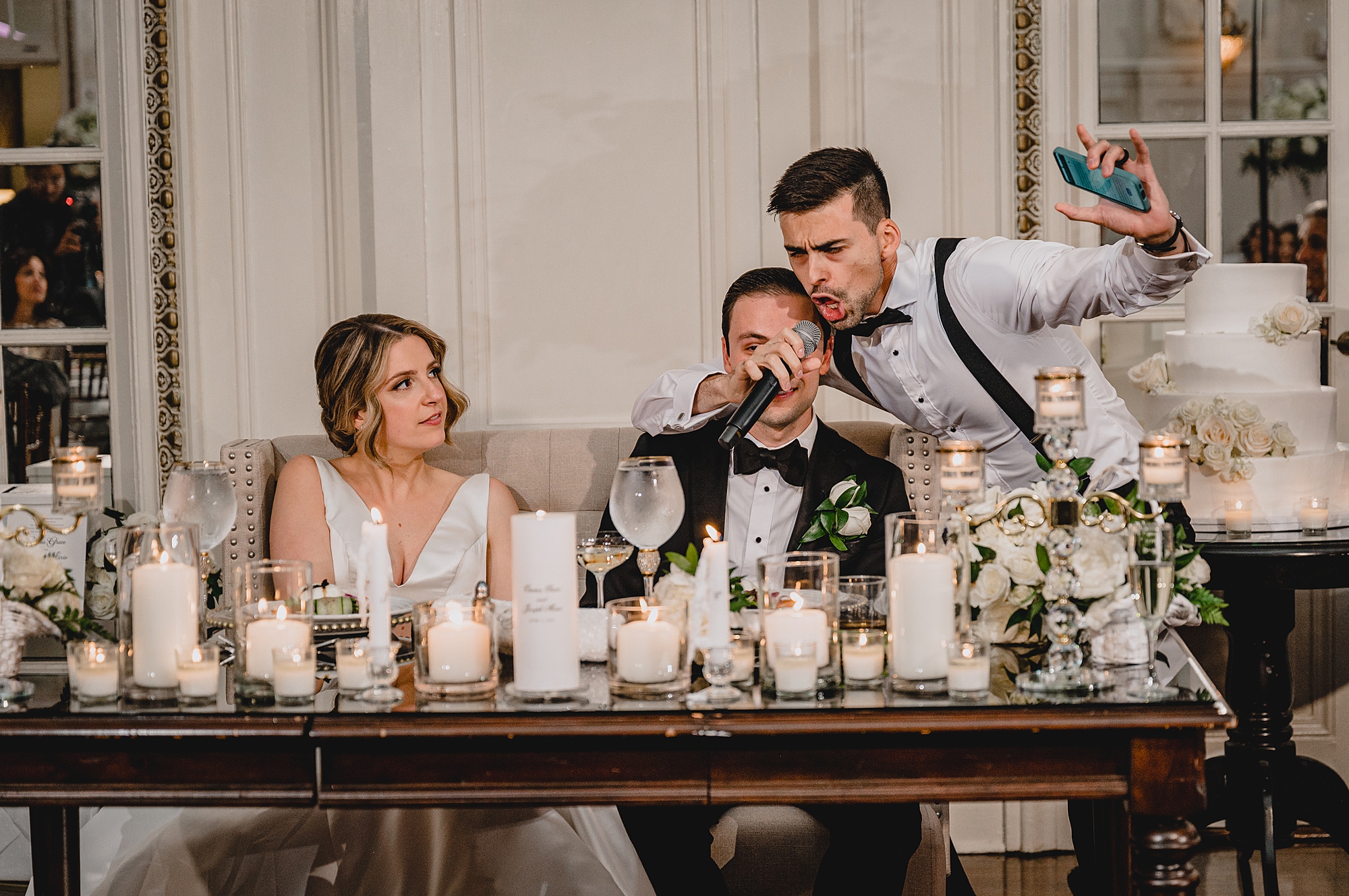 groomsman hugs groom at sweetheart table during Long Island wedding reception in the ballroom