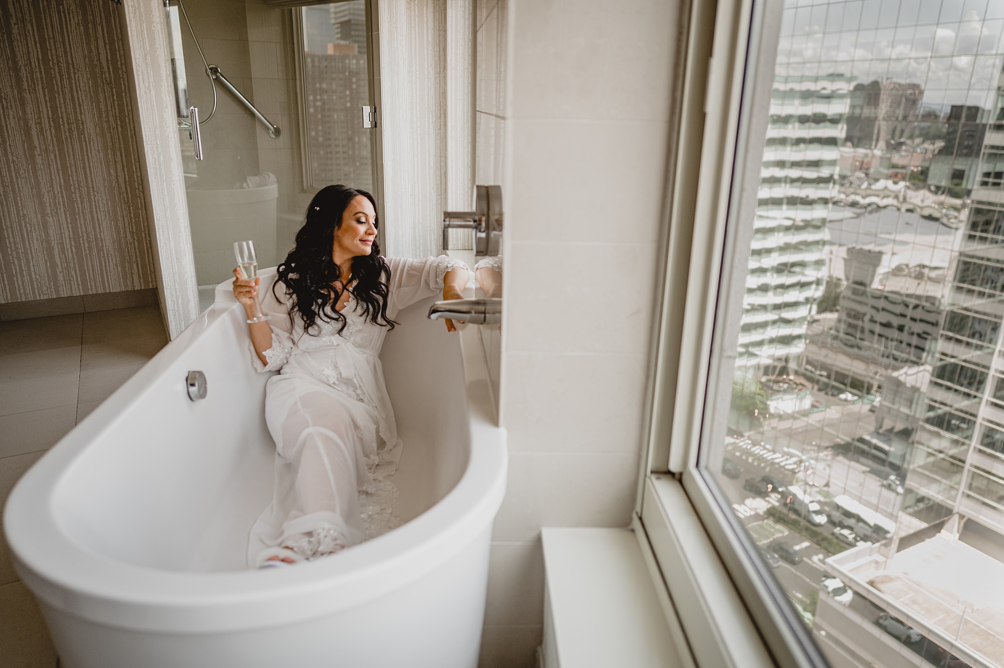 bride sits in white pajamas in bathtub of hotel room