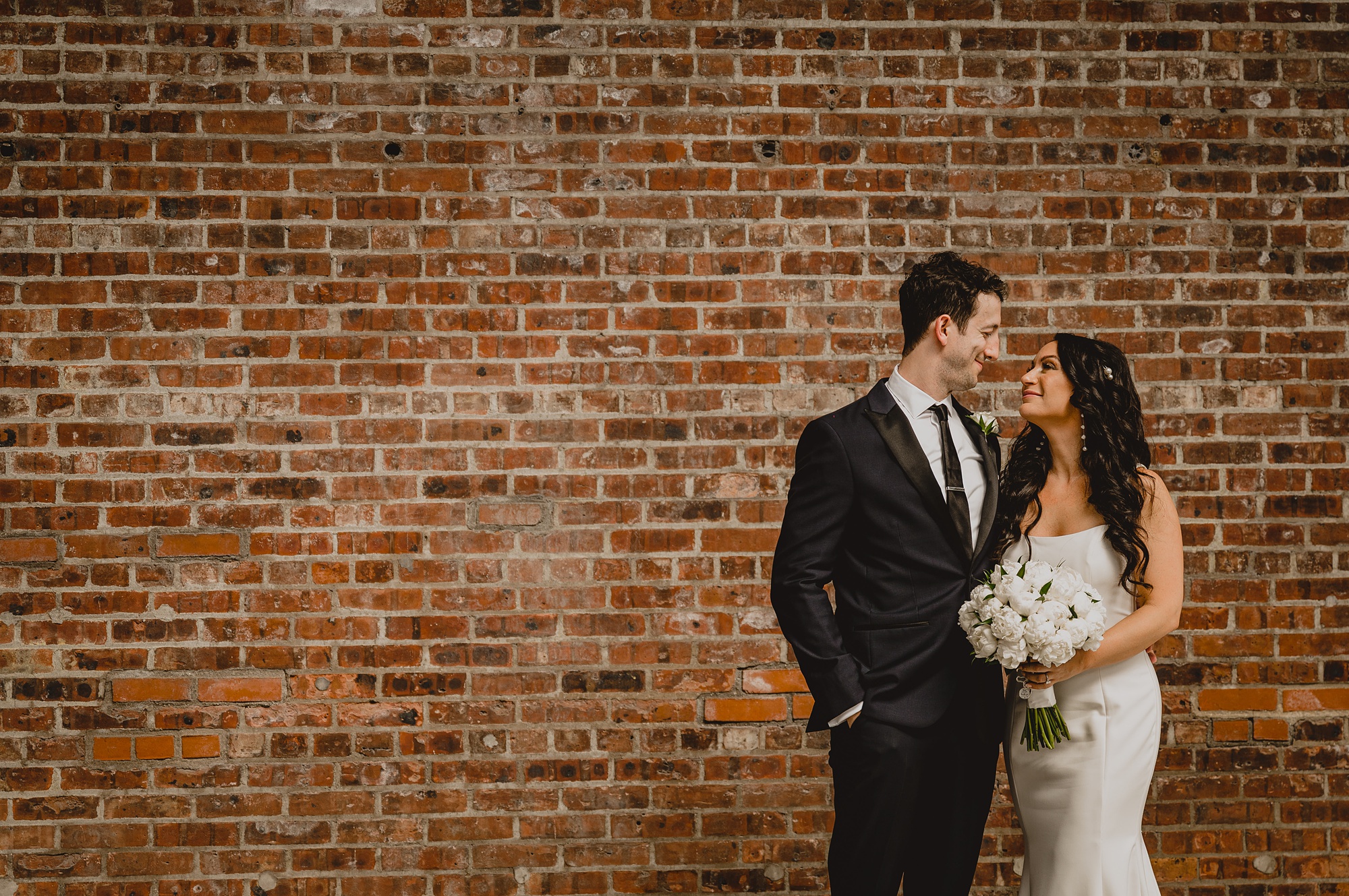 groom smiles at bride during portraits by brick wall at the Liberty House