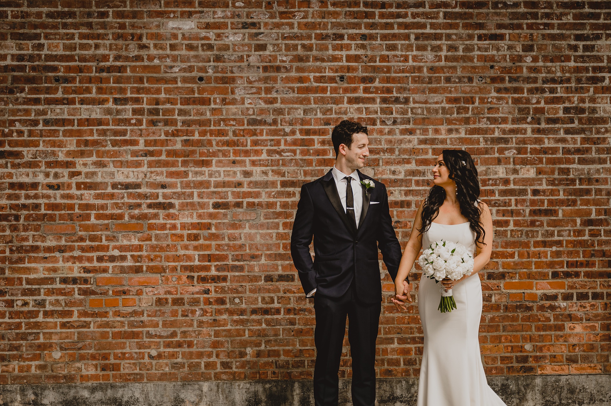 bride and groom hold hands standing by brick wall at the Liberty House