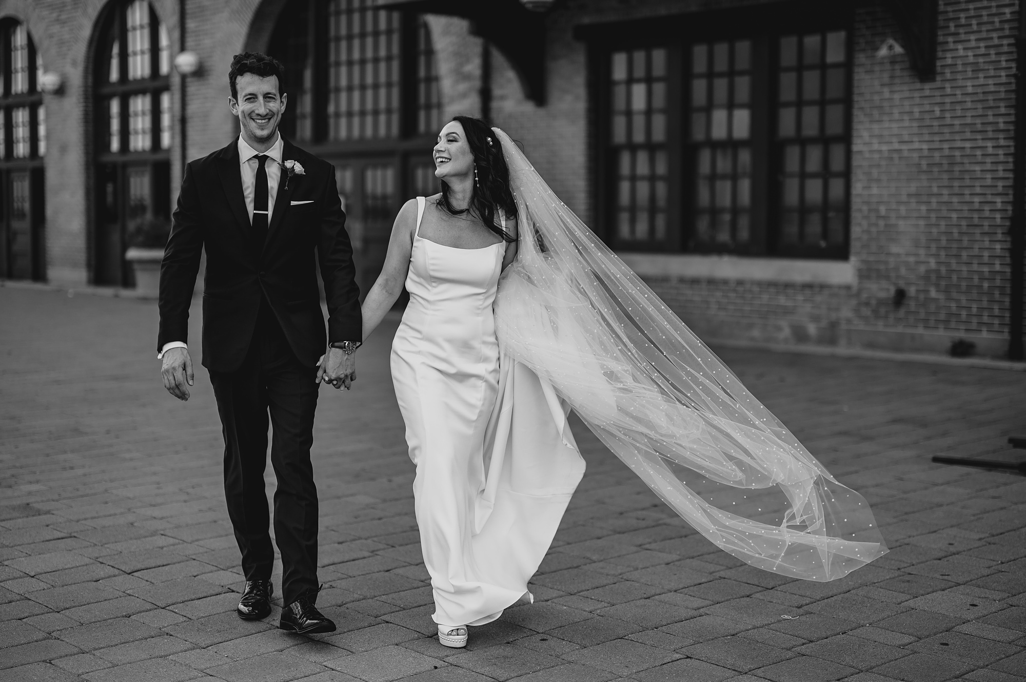 bride and groom hold hand walking outside the Liberty House with veil floating in air