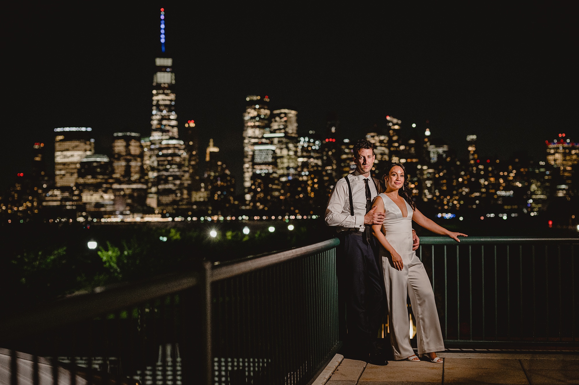 newlyweds hug leaning against railing in front of the Hudson River with NYC skyline behind them at night