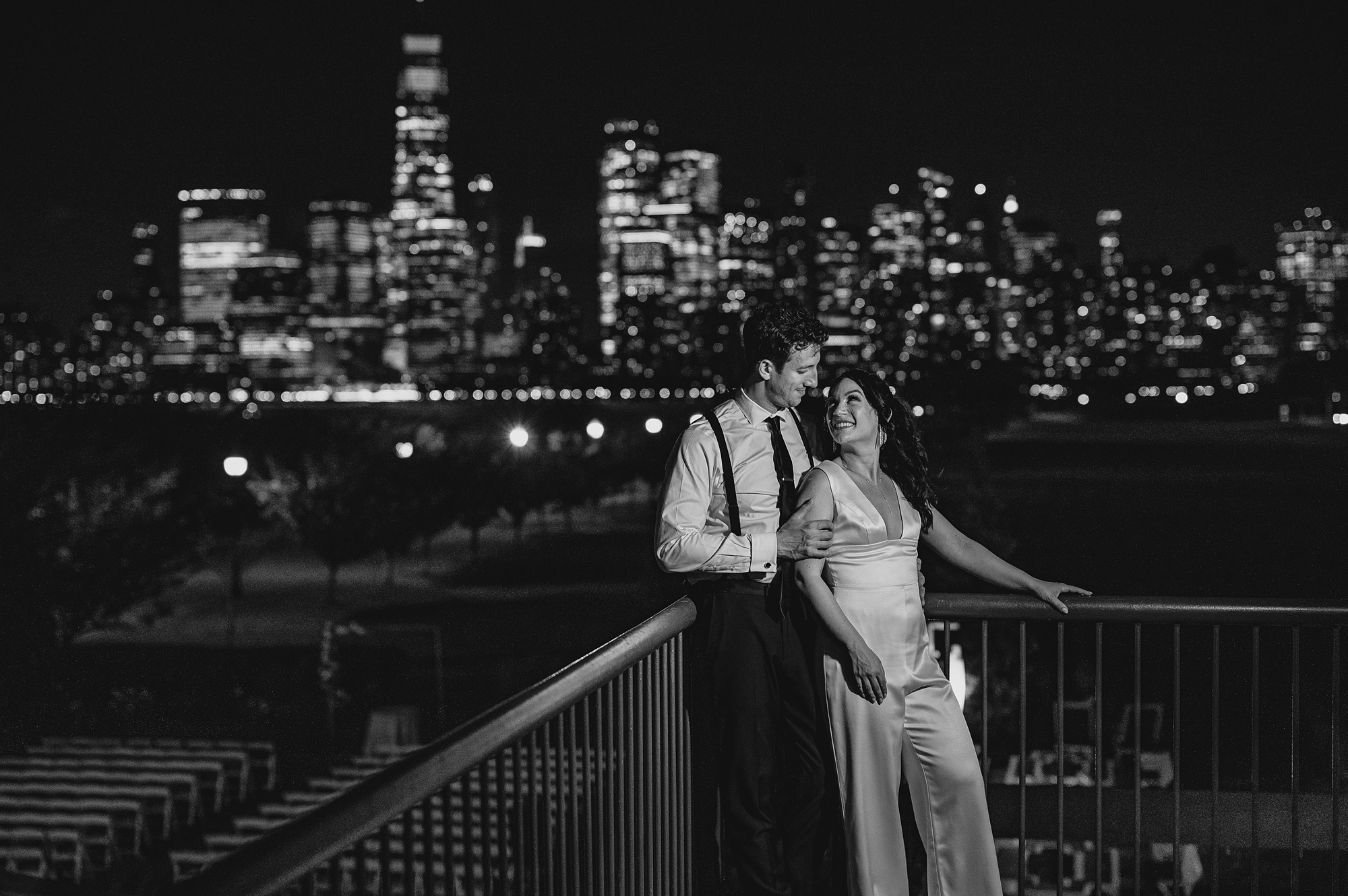 newlyweds hug leaning against railing in front of the Hudson River with NYC skyline behind them at night
