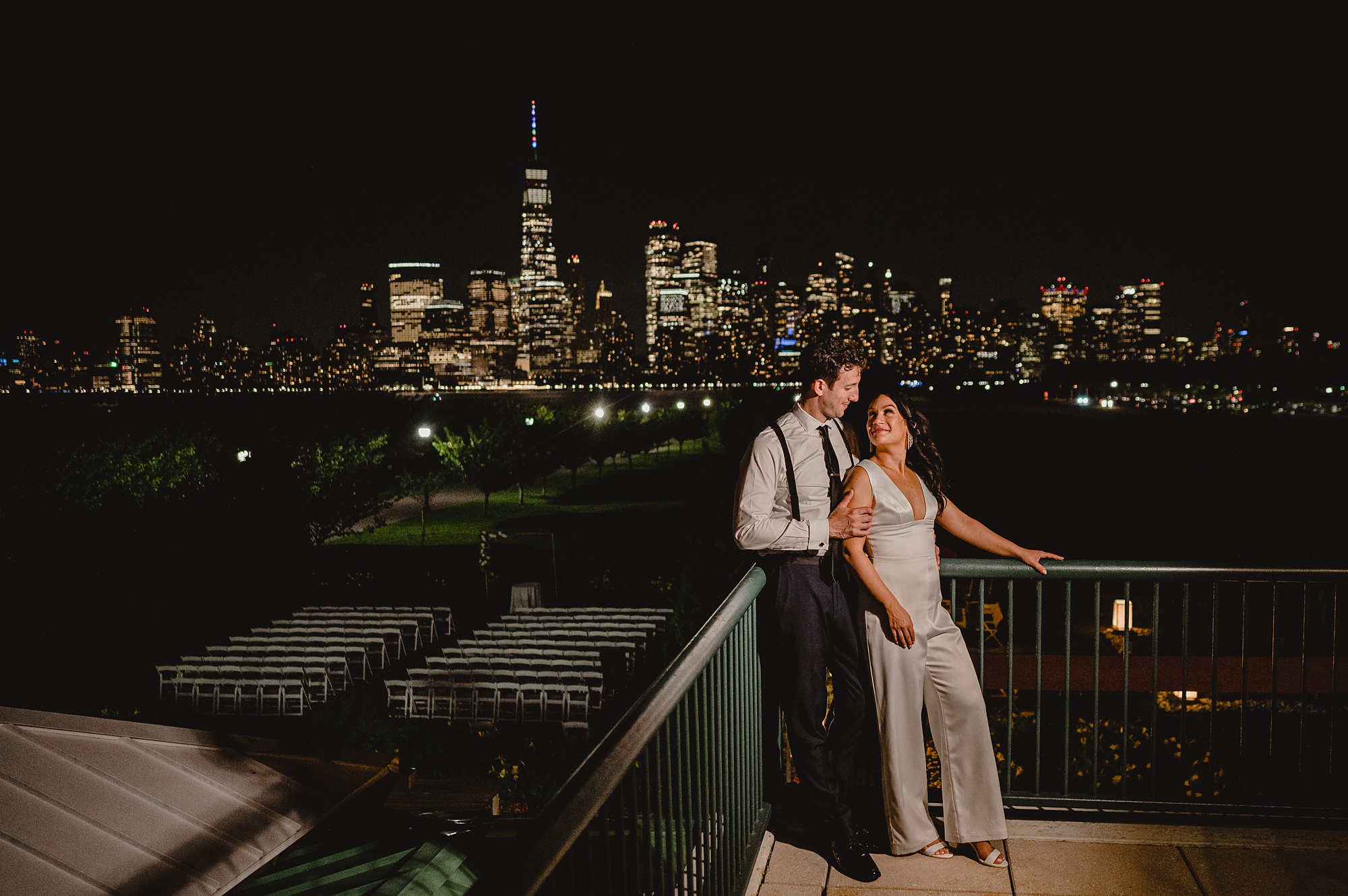 newlyweds hug leaning against railing in front of the Hudson River with NYC skyline behind them at night