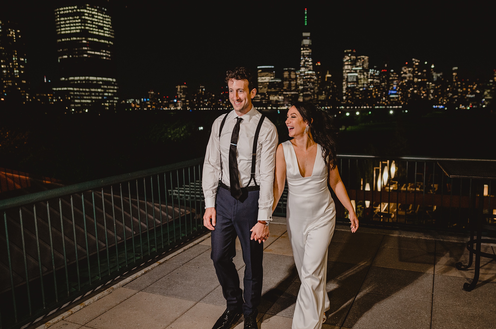 bride and groom walk across patio in front of the Hudson River with NYC skyline behind them at night