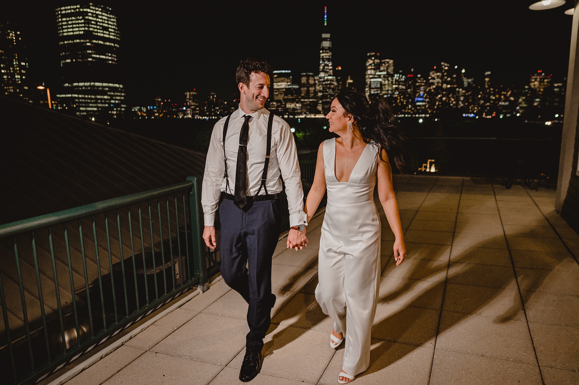 bride and groom walk across patio in front of the Hudson River with NYC skyline behind them at night