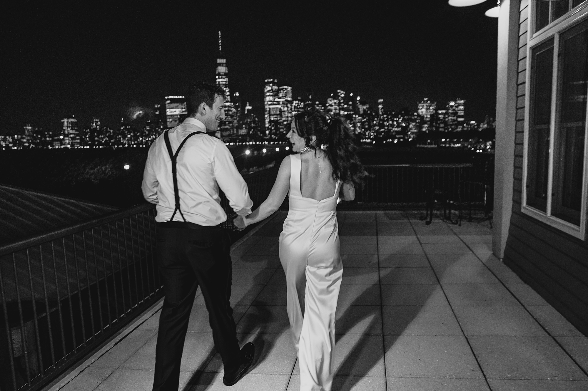 bride and groom walk across patio in front of the Hudson River with NYC skyline behind them at night
