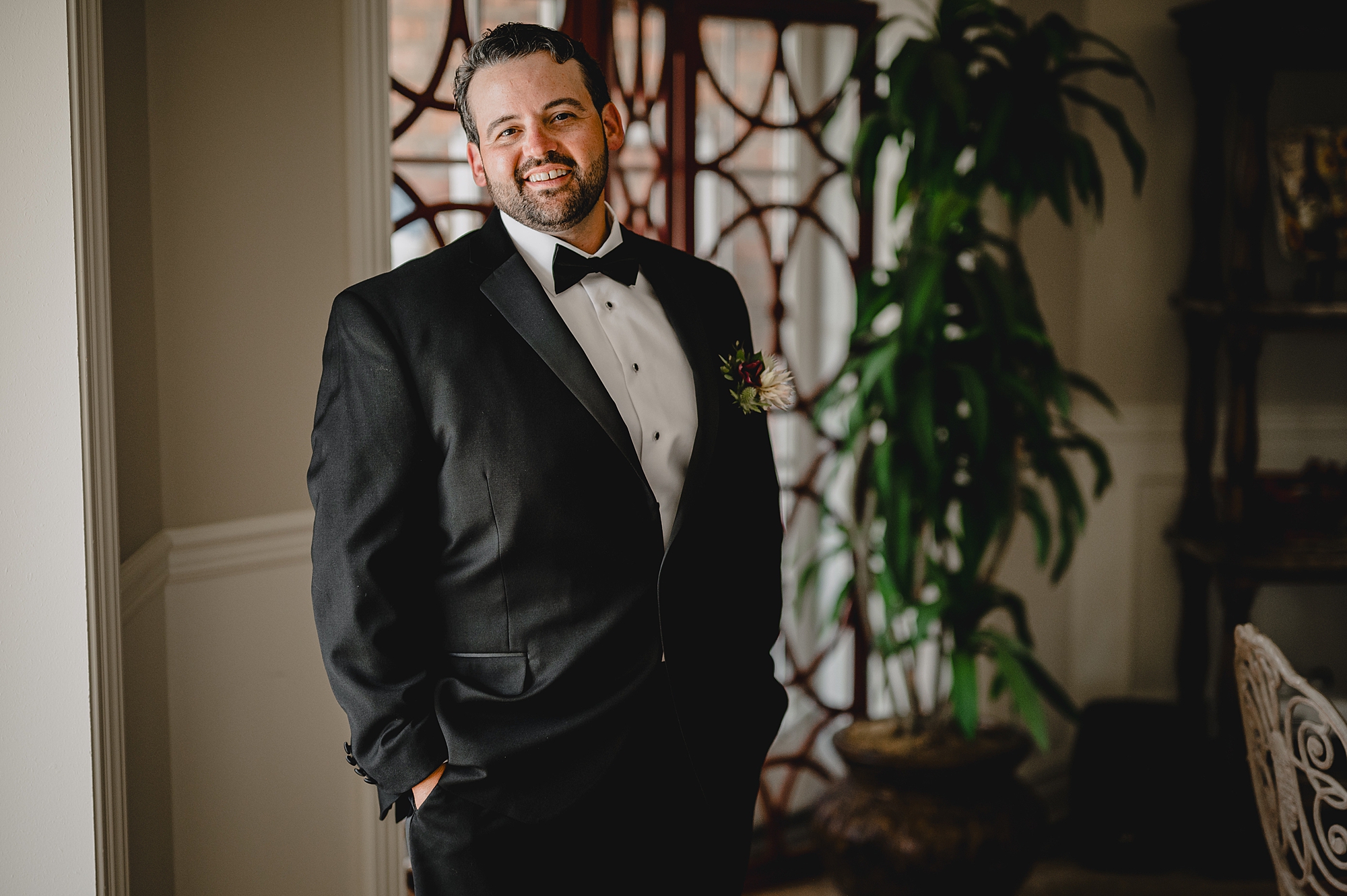 groom stands with hands in pockets of suit smiling in front of window