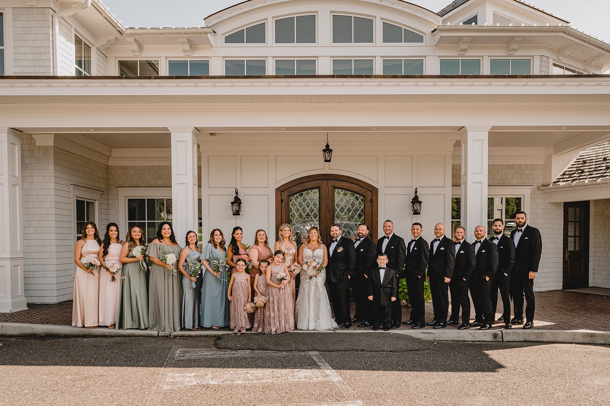 bride and groom stand on front porch of the Mill Lakeside Manor with wedding party in black tuxes and pink dresses