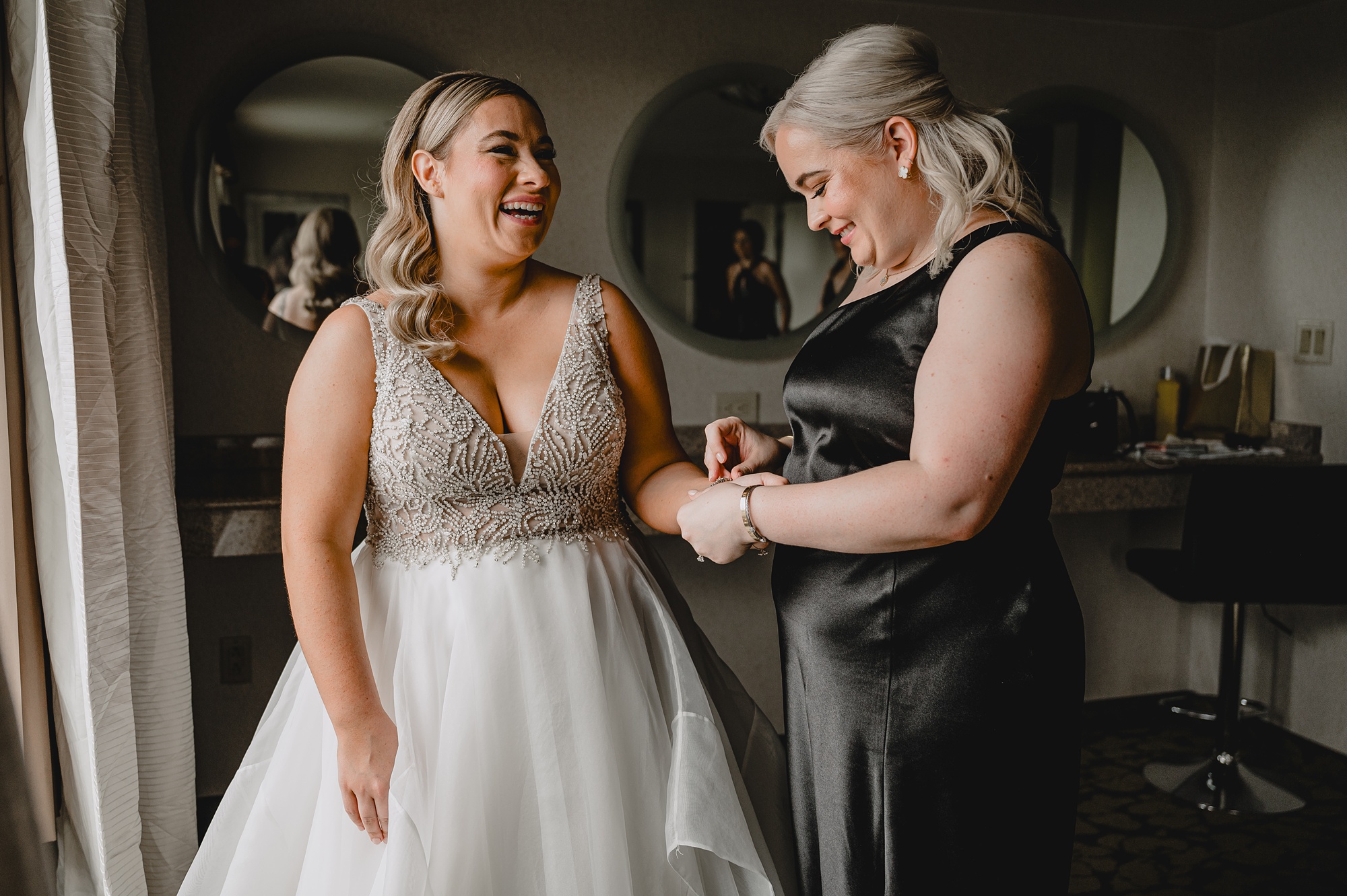 bridesmaid in black gown puts on jewelry for bride in Staten Island hotel