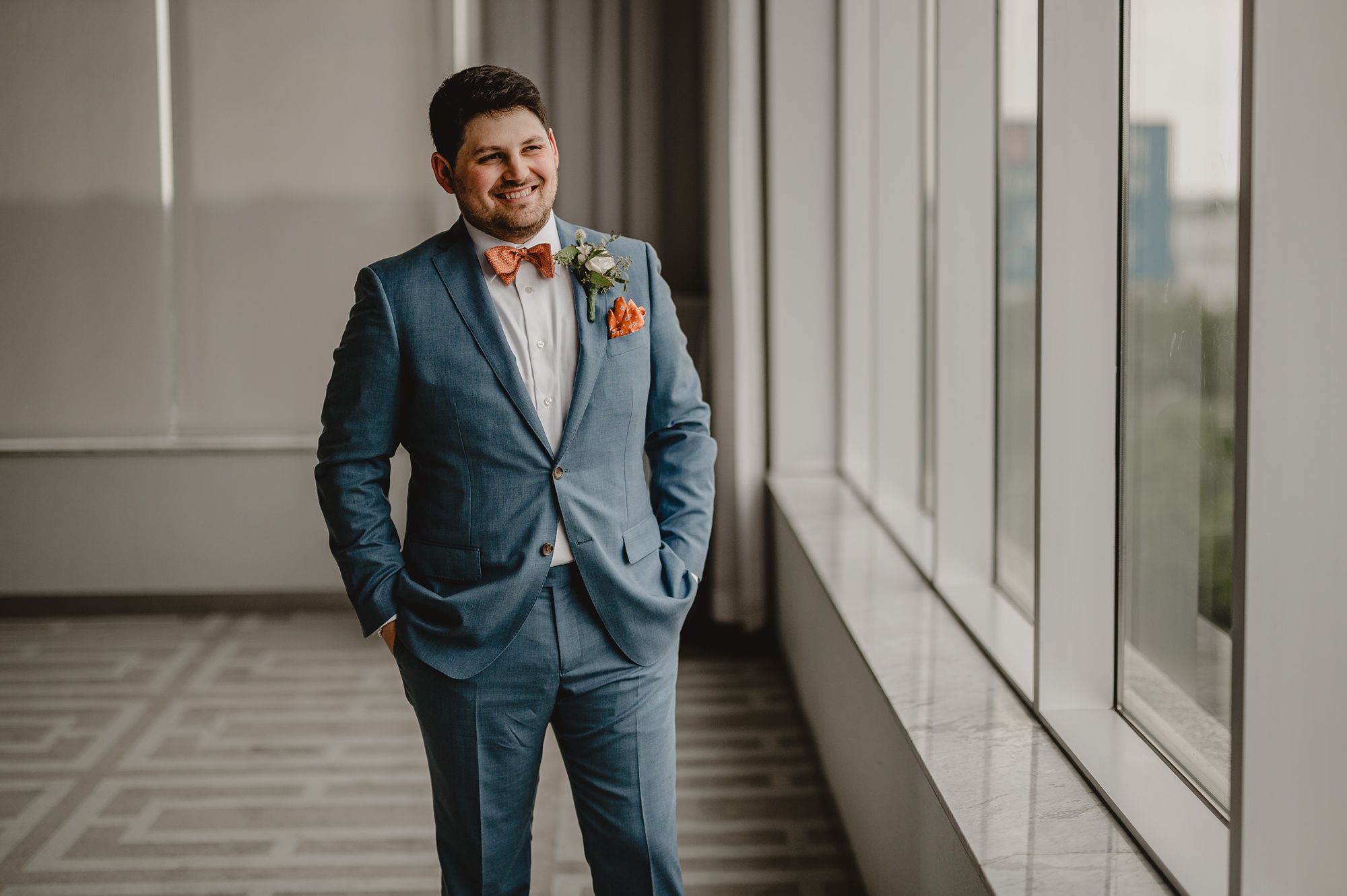 groom stands by window of Above Staten Island in blue suit