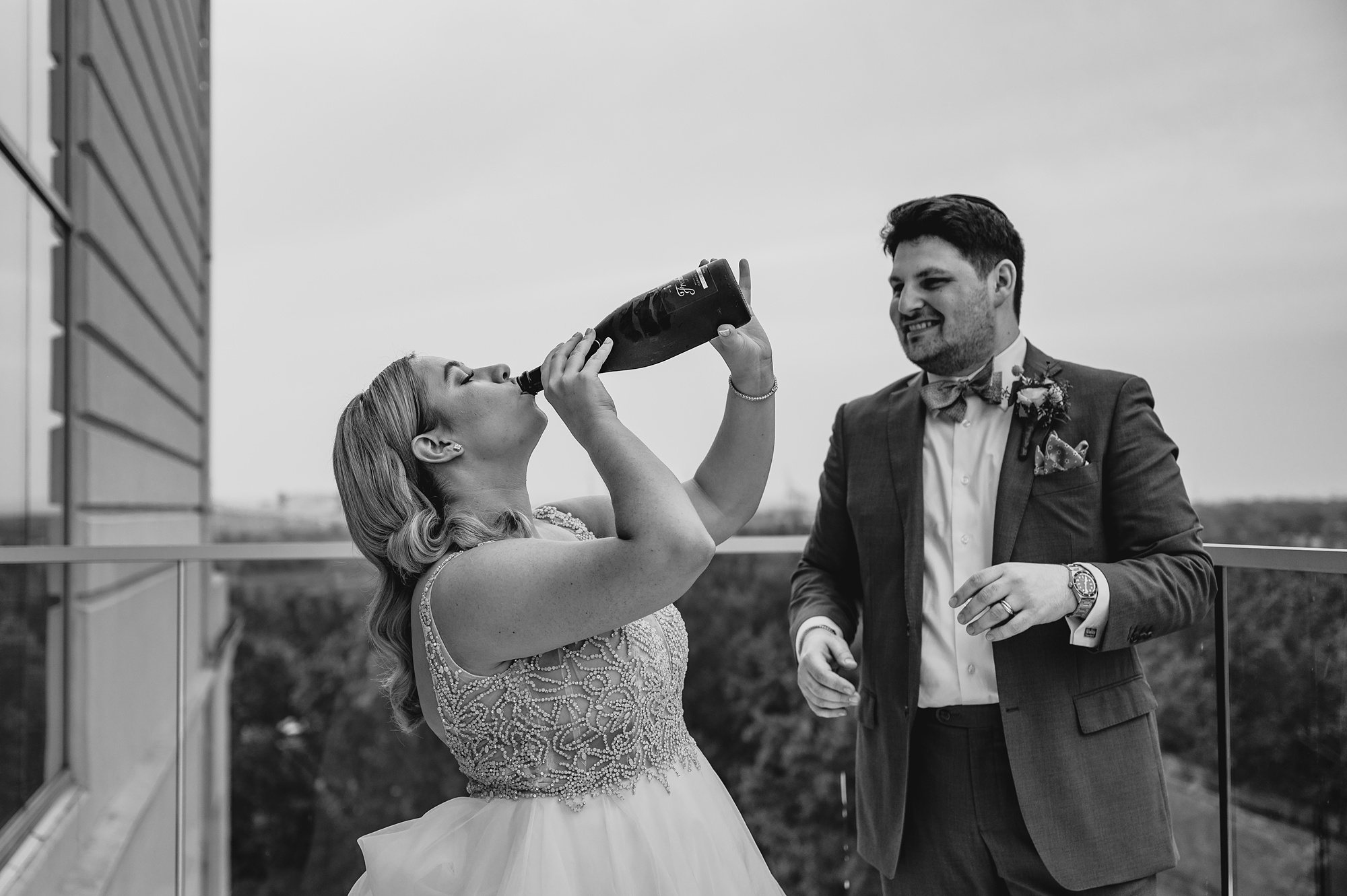 bride drinks champagne from bottle while groom watches 