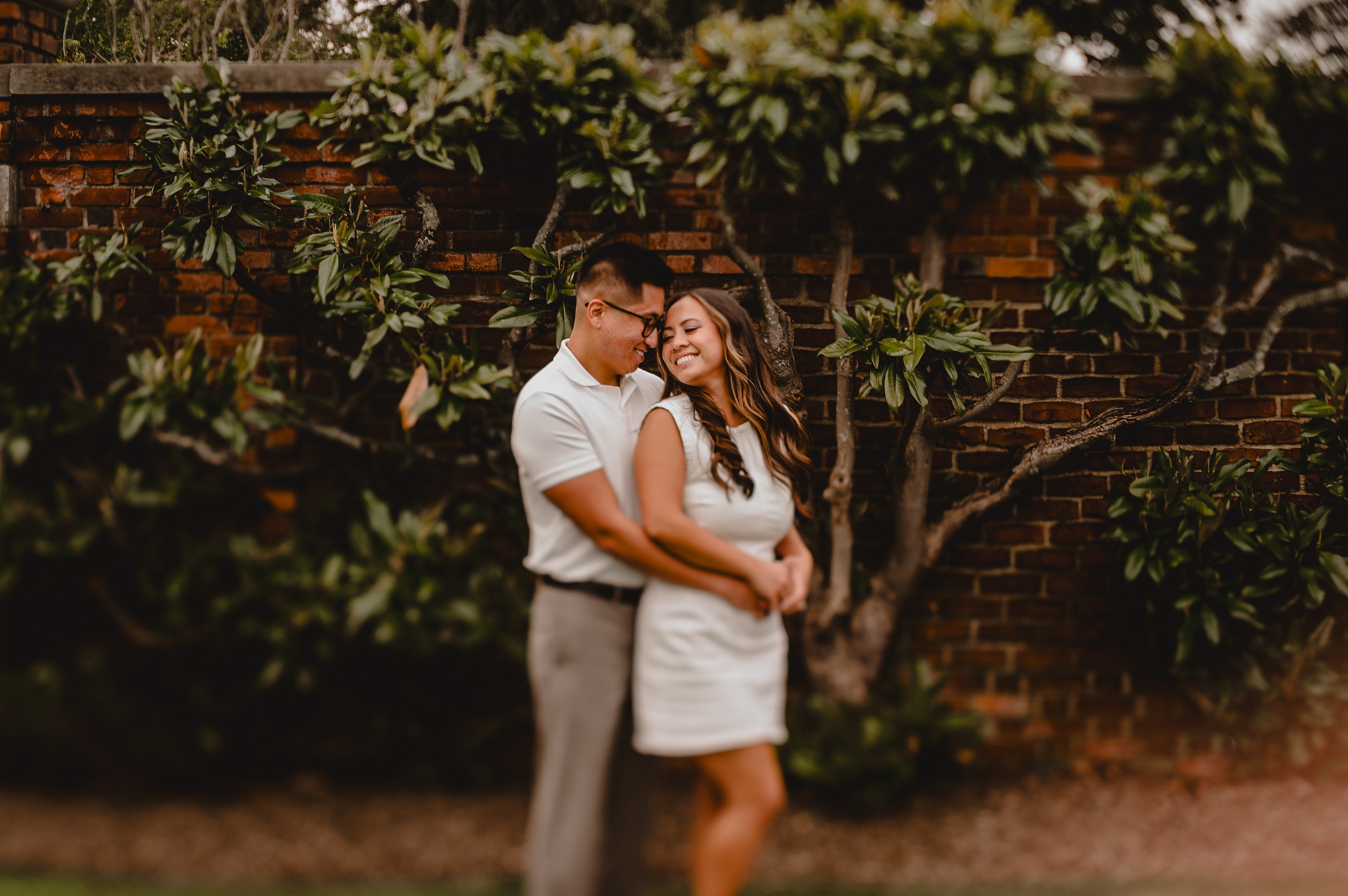 man hugs woman around waist in front of tree and brick wall at Old Westbury Gardens