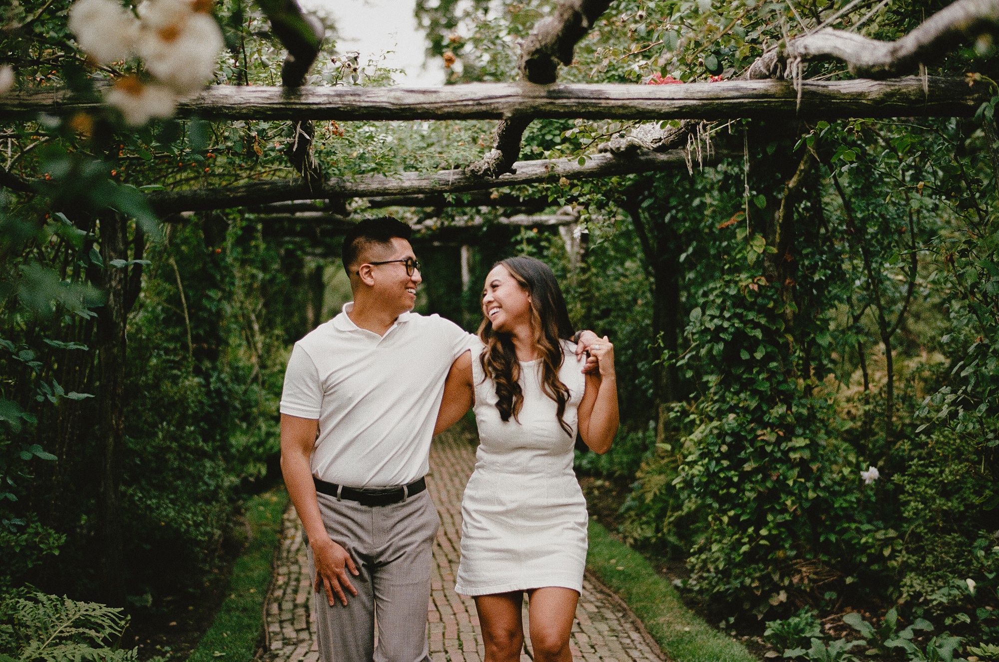 engaged couple walks under arbor holding hands on each other's shoulders 