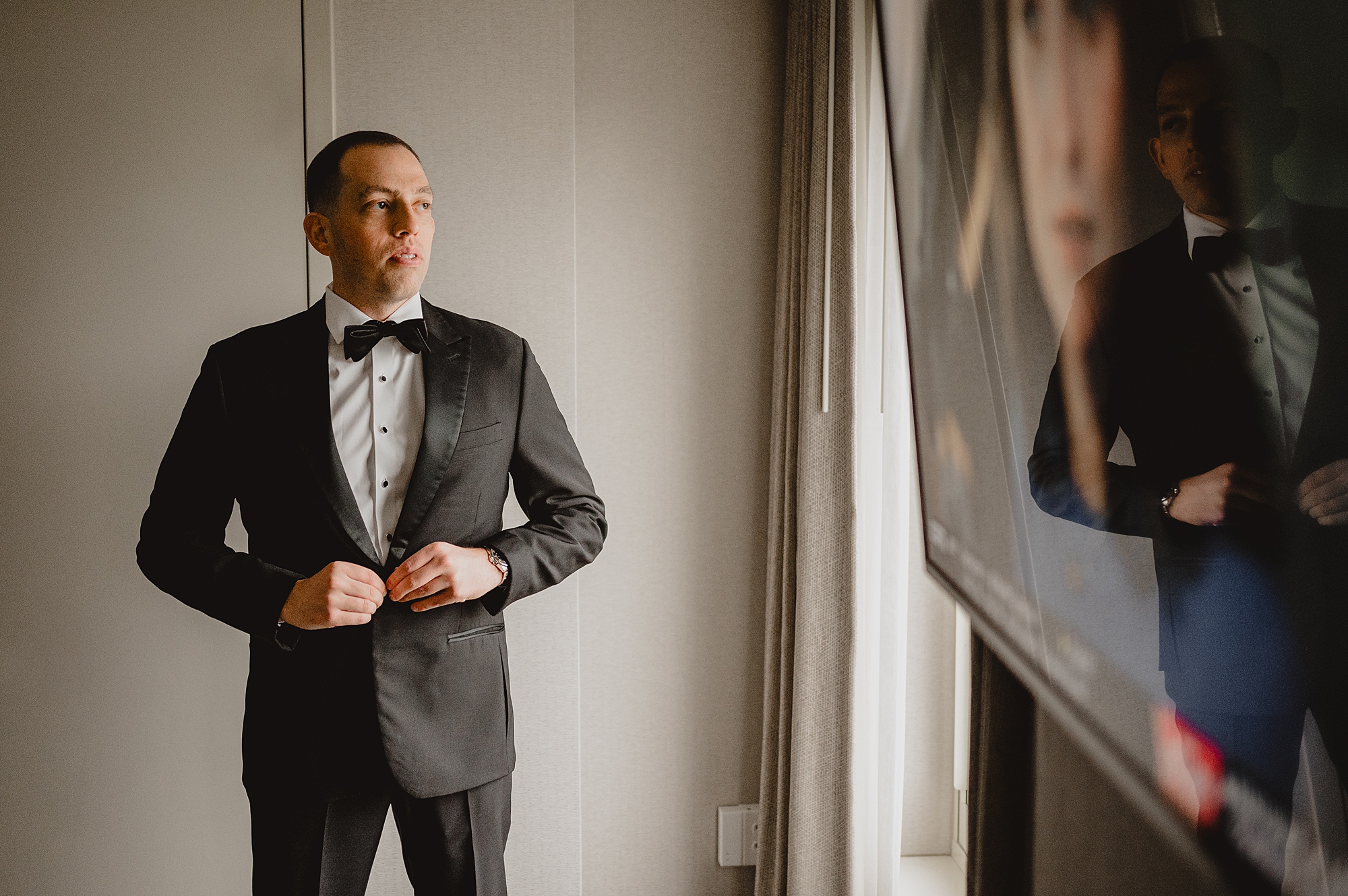 groom buttons up tux jacket in window of Jersey City hotel room