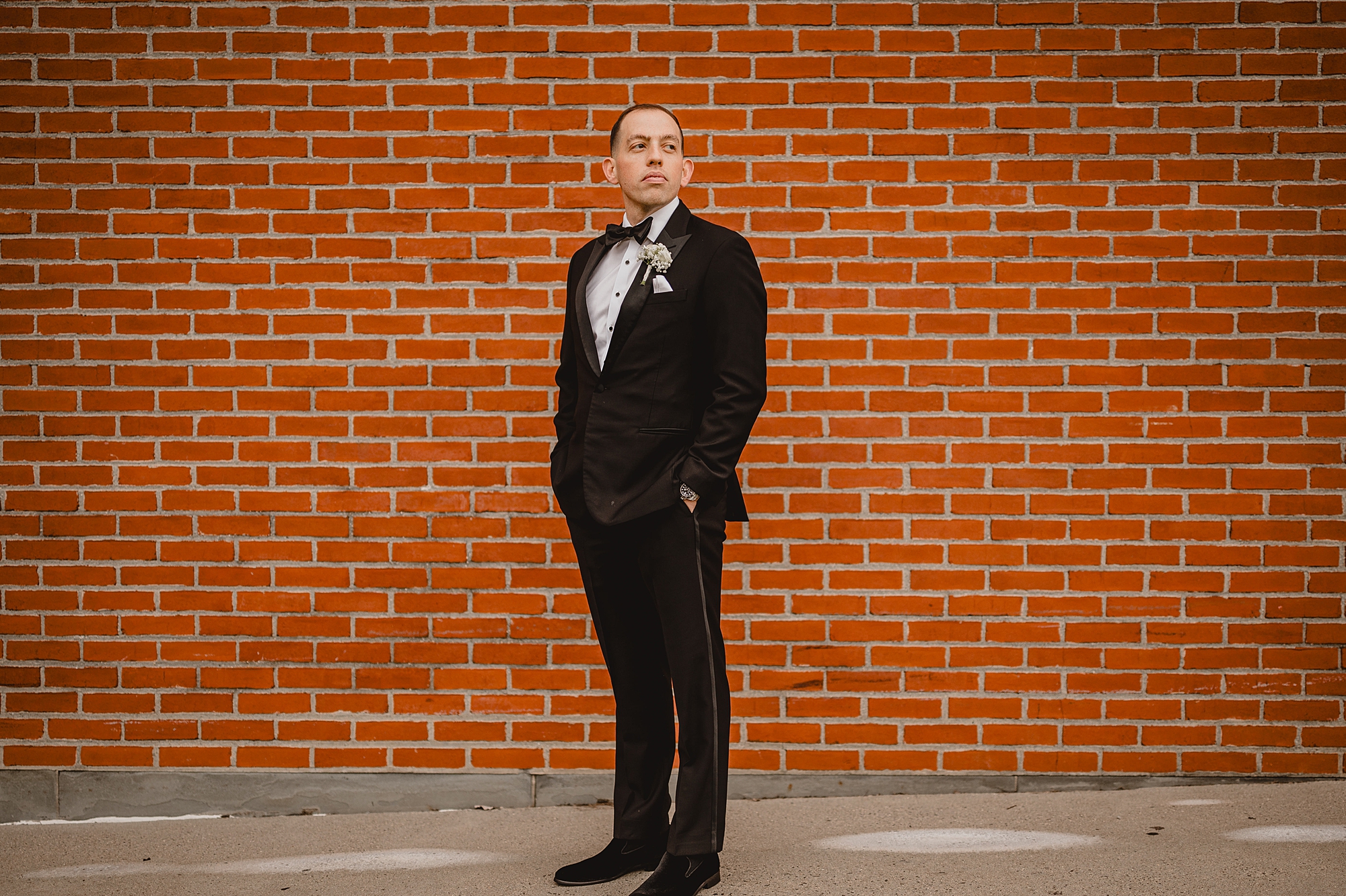 groom stands with hands in pockets in front of brick wall at the Liberty House