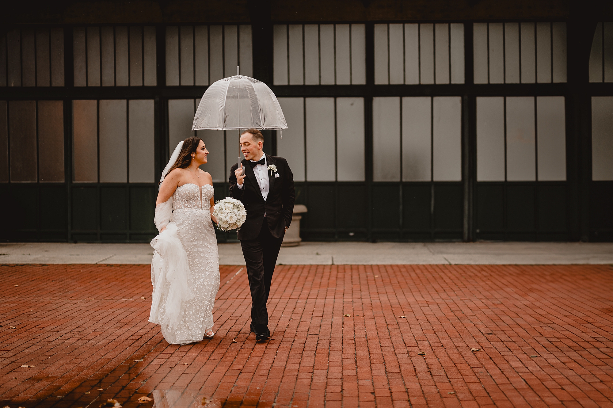 newlyweds hold hands walking under clear umbrella outside the Liberty House