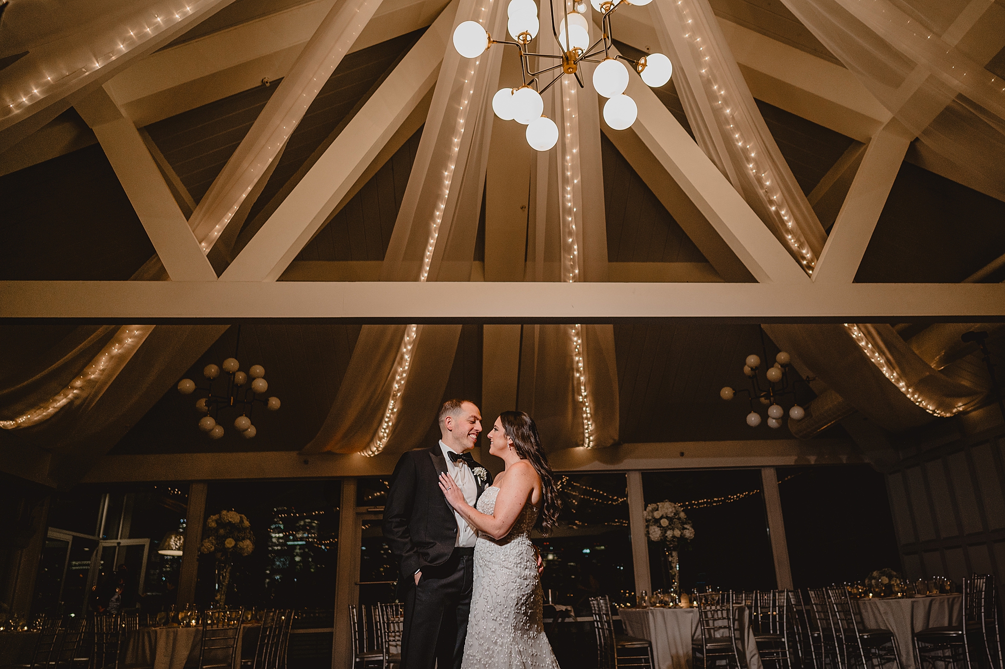bride and groom hug under white rafters lined with lights