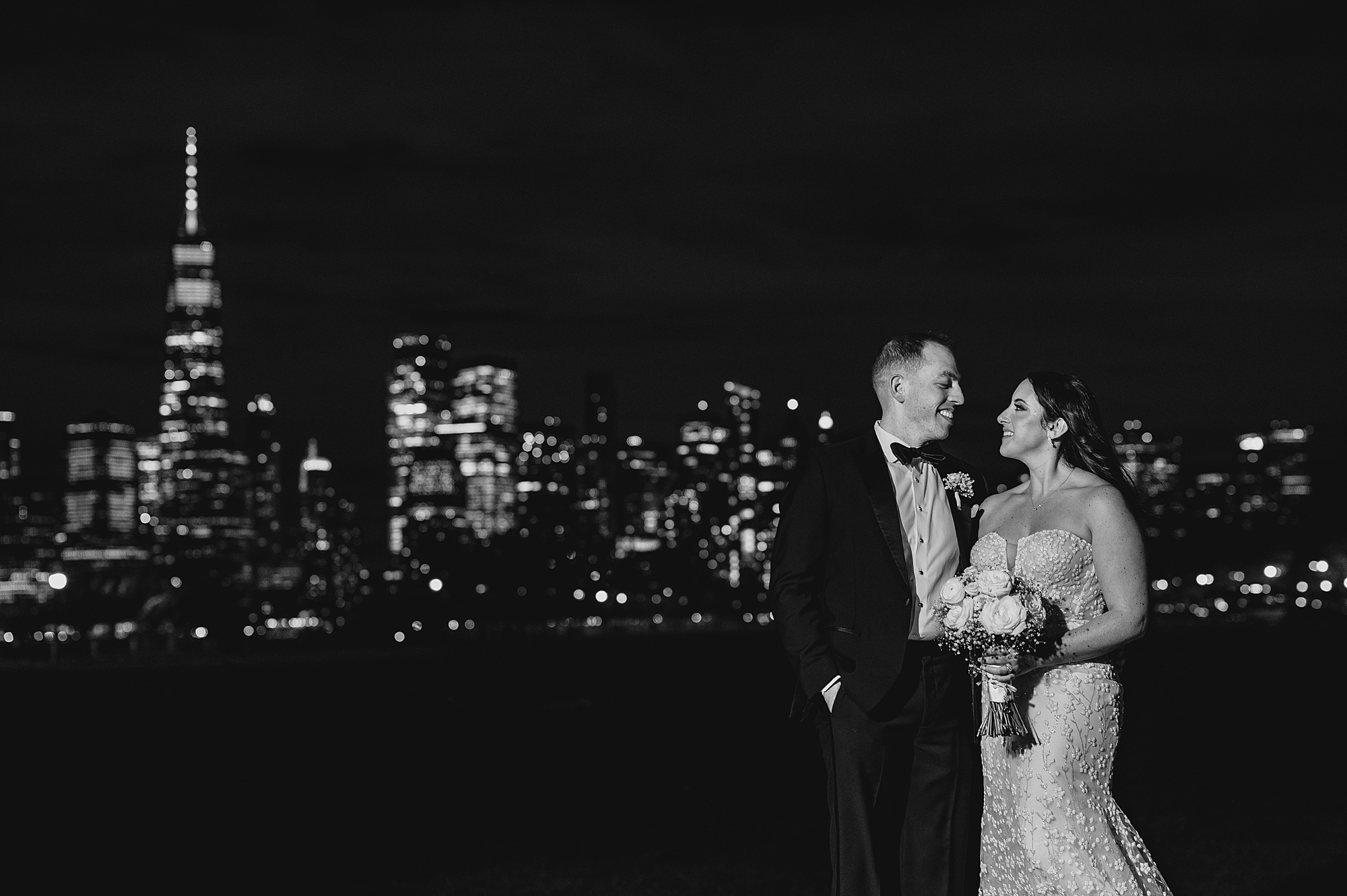 newlyweds hug on patio at night with NYC skyline lit up behind them