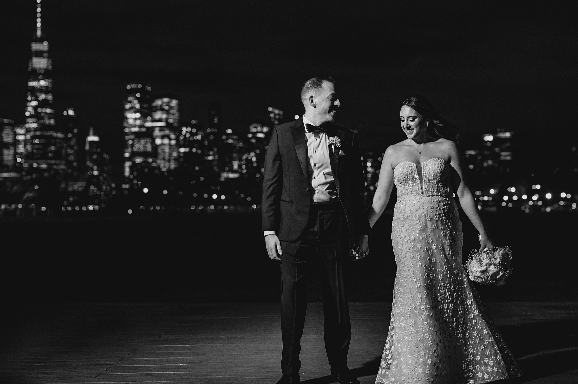newlyweds hold hands in front of lit NYC skyline at the Liberty House